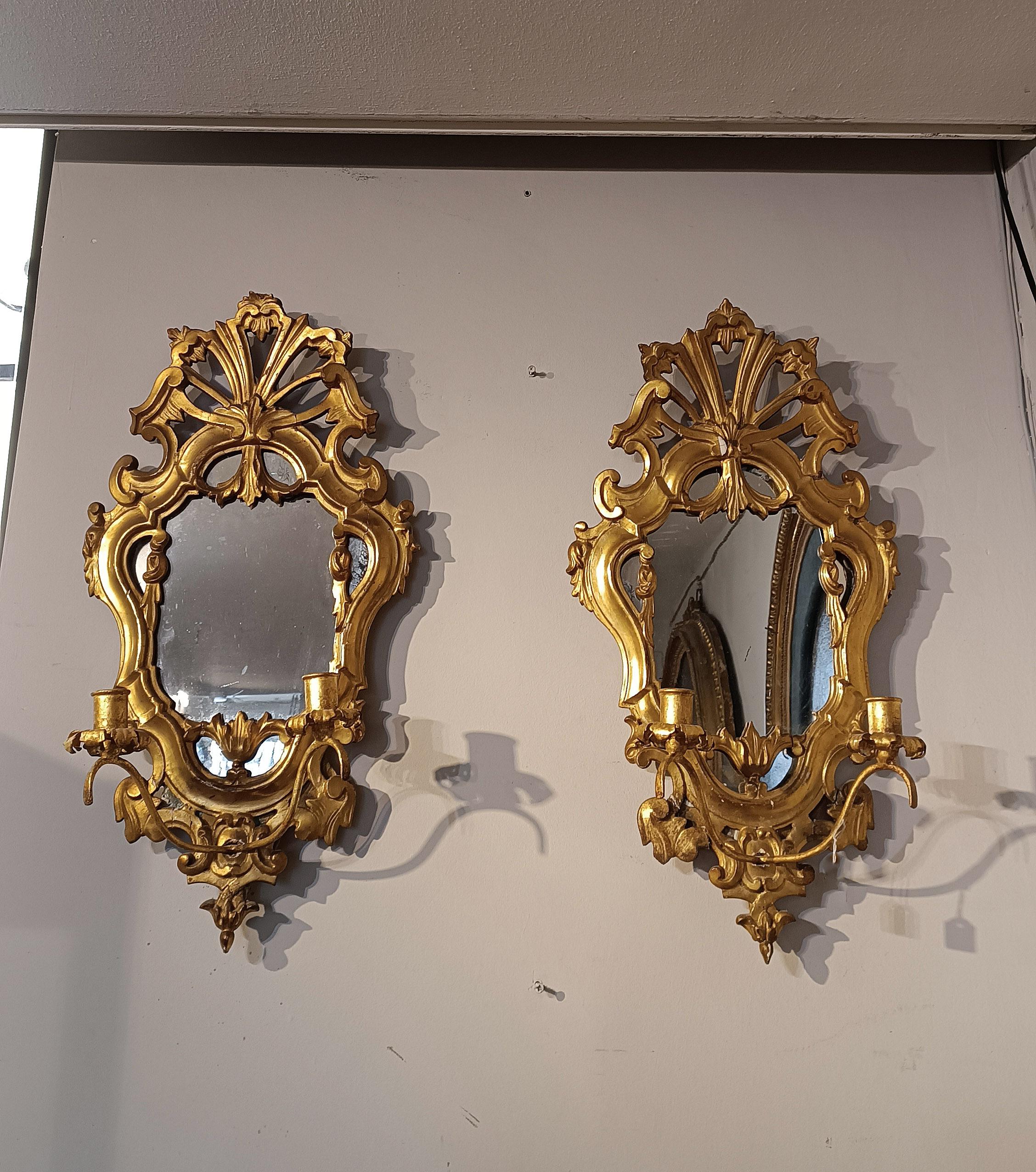 Elegant pair of small mirrors in pine wood, finely carved and gilded with pure gold leaf. The frames are decorated with scrolls and vegetal motifs, as is the cymatium which features an elaborate curl. The mirrors are equipped with candle holder arms