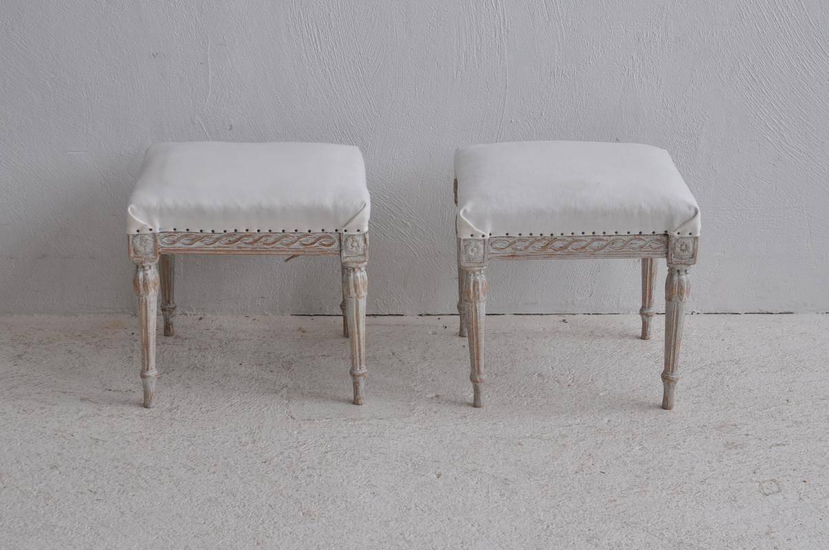 A pair of Swedish wood stools with upholstered seats from the Gustavian period. These footstools are covered in similar, but different fabrics. There is a carved guilloche pattern on the seat frame with corner blocks featuring carved rosettes above