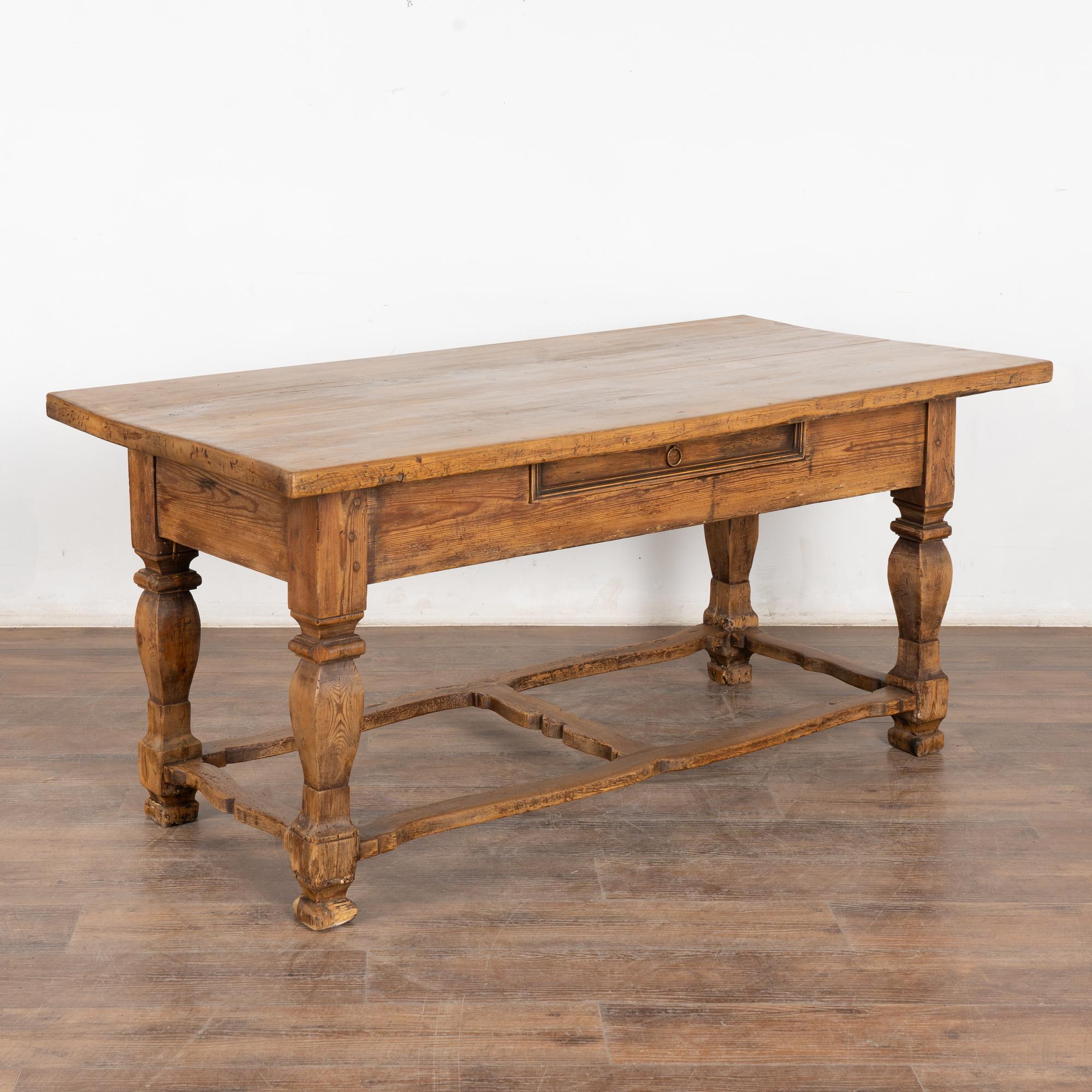This handsome pine library table with one drawer has heavily turned legs, and unique carved/curved stretchers.
The top and legs reveals nicks(see feet), gouges, cracks and stains reflecting generations of use. 
Restored, stable, drawer functions.