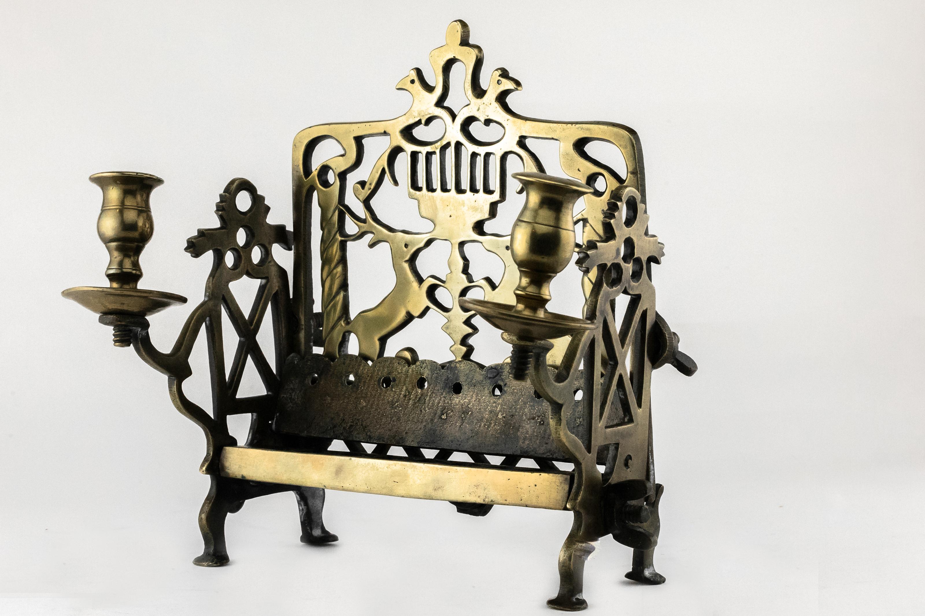 Chanukka-Lampe aus Messing, Polen, 18. Jahrhundert.
Messing, gegossen, ein durchbrochener, rechteckiger Sockel ist an den Seiten, die auf vier Beinen stehen, und an der Rückwand angeschraubt. Quaderförmiges Tablett, innen in acht würfelförmige