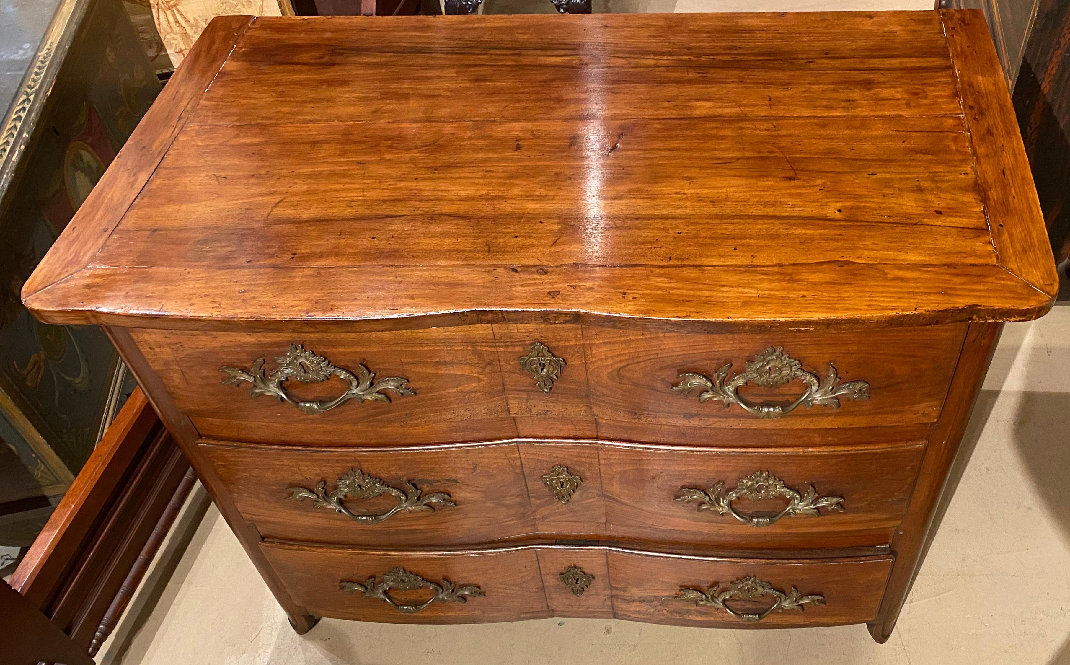 A fine provincial walnut three drawer commode with shaped top and conforming case, scrolled brass or bronze pulls and escutcheons (probably original), raised panel drawer fronts and side panels, all supported by two front short splayed carved feet.