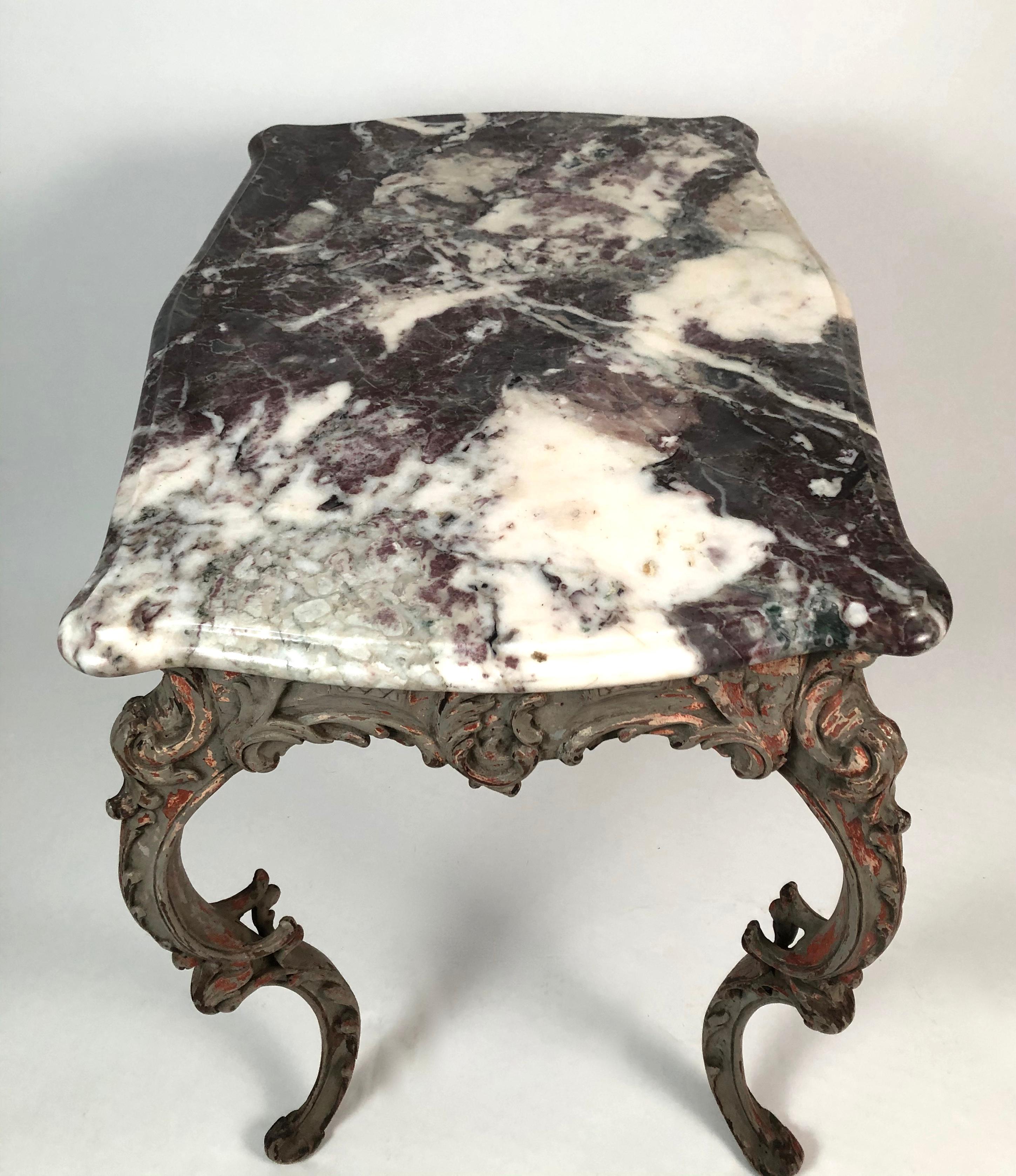 An 18th century Italian Rococo table with a serpentine breccia violetta--a variegated lilac colored marble-- top over a floral carved apron and boldly curved C-scrolled legs, in grey paint which shows coral pigment underneath from when it may have