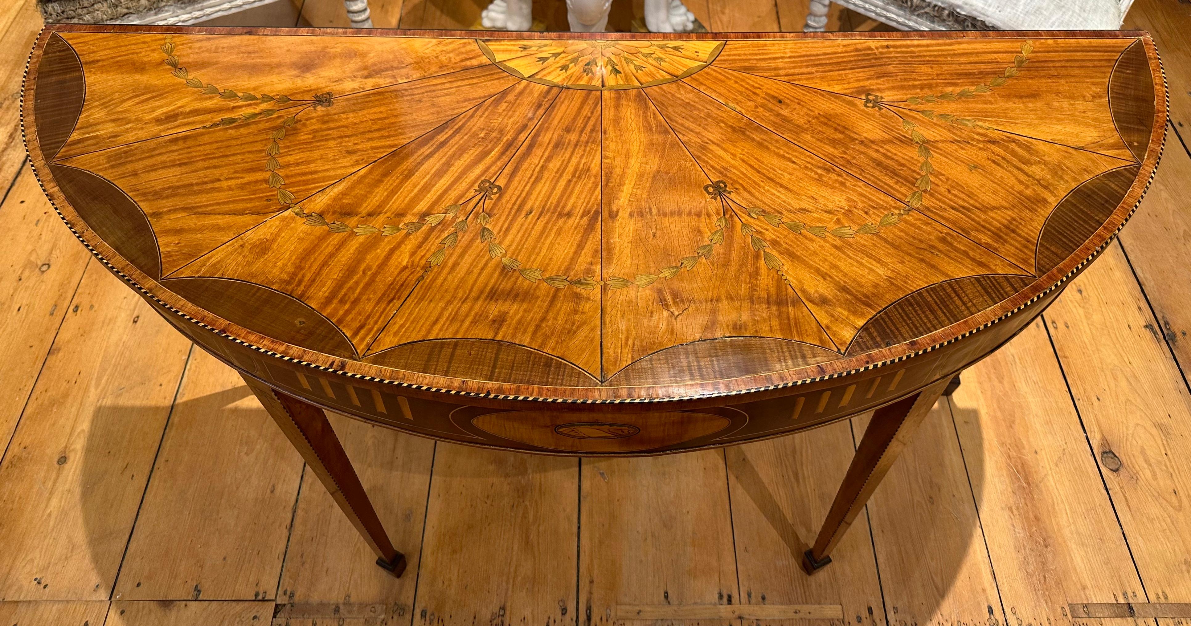 George III marquetry veneered demilune table, ca 1785. The top and sides with satinwood and harewood veneers and stringing, the center frieze with an inlaid and stained shell and flutes, above similarly inlaid tapering square legs.  Inlaid