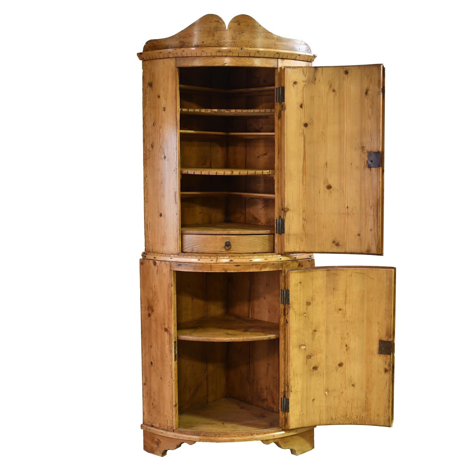 A charming Baroque corner cupboard with arched pediment top and two bowed doors with original steel hinges that offer two separate storage spaces that open to shelving with one interior drawer. Base rests on bracket feet with scalloped edge. Comes