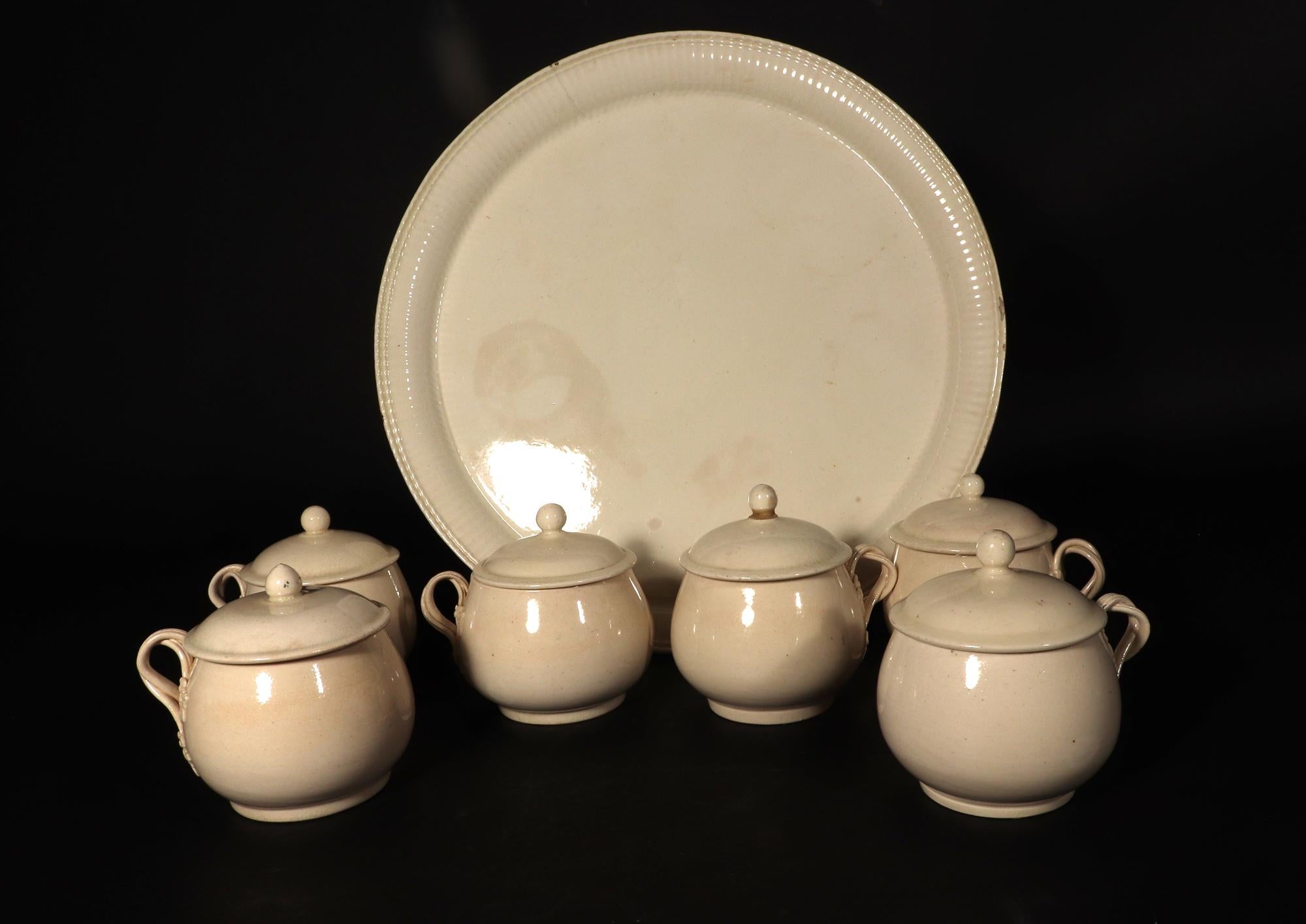 Juego de Pot de Creme inglés y soporte,
Juego de seis,
Hacia 1780-85

Las vasijas de cerámica cremosa tienen forma de bombé circular con un remate de botón circular liso.  Las asas son de doble lazo con forma de tiras reed con terminales de hoja. 