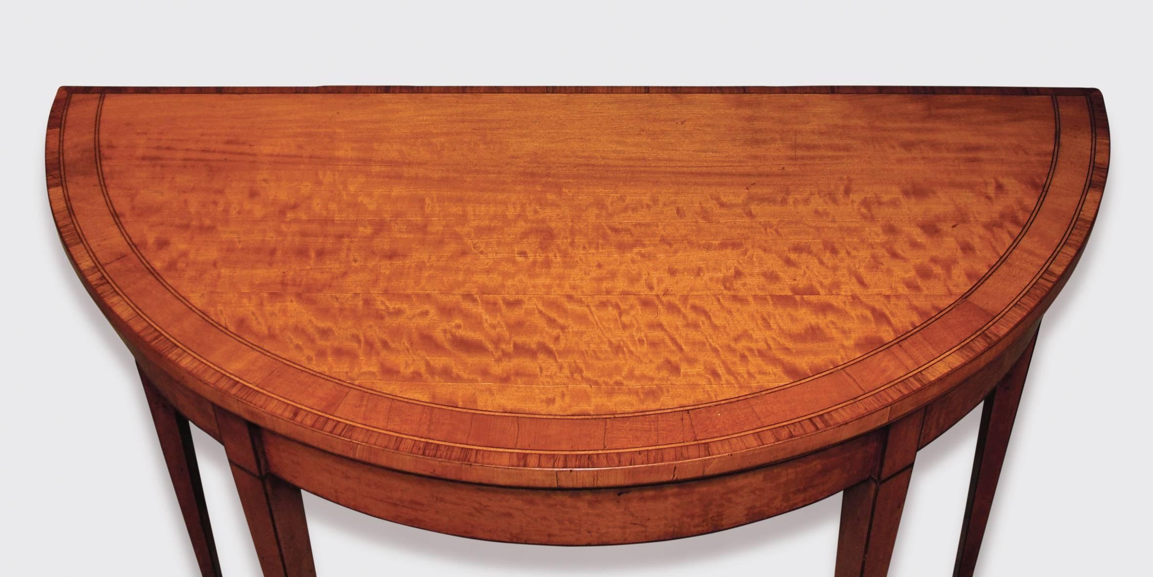 A late 18th century Sheraton period satinwood card table, boxwood and ebony strung throughout, having satinwood and tulip wood crossbanded demilune top, above frieze, supported on square tapering legs.