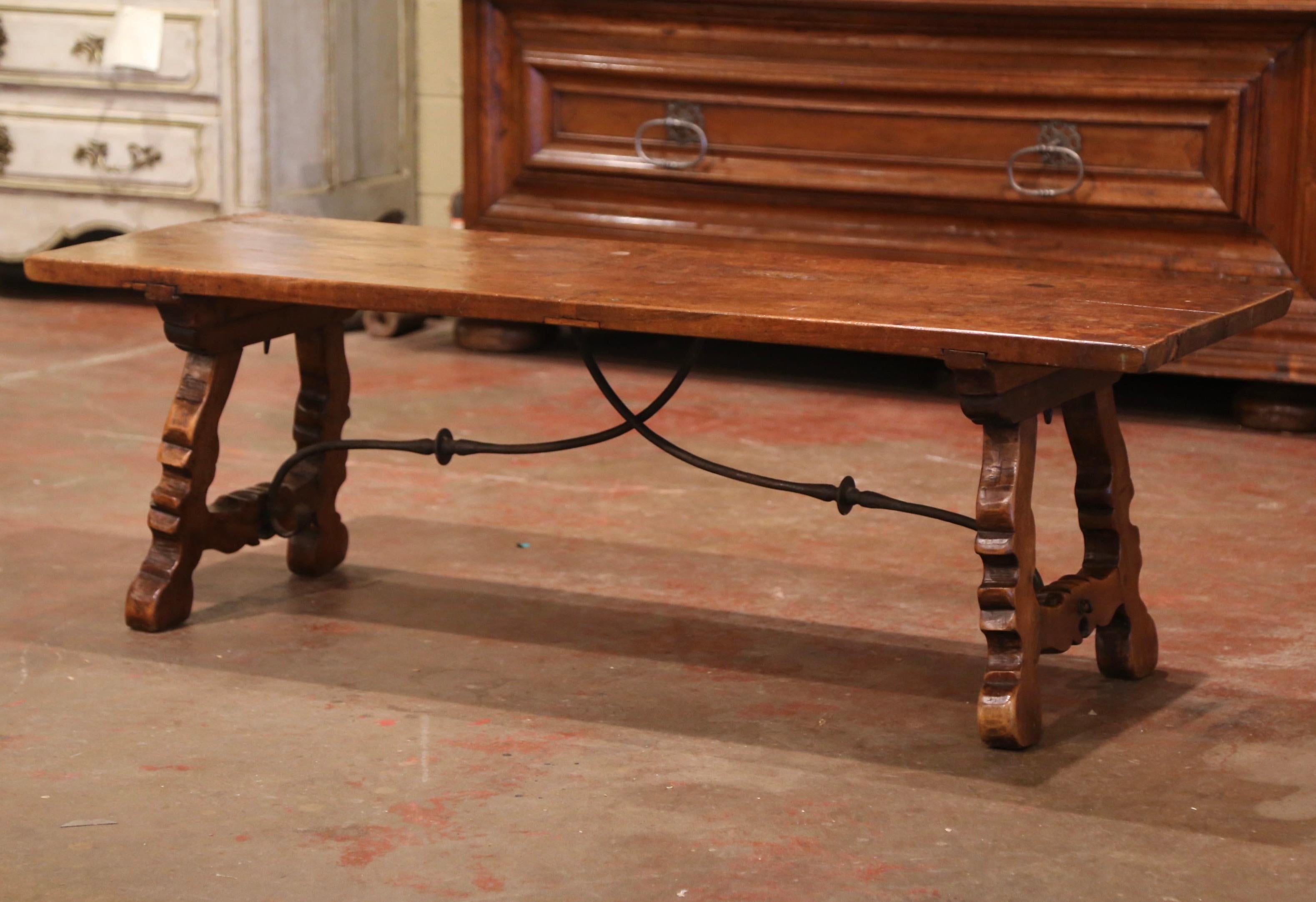 Crafted in Spain circa 1780, the rectangular antique fruit wood coffee table stands on two carved scrolled legs joined together with a forged iron stretcher; the top is built with a single plank of walnut. The long and narrow cocktail table is in