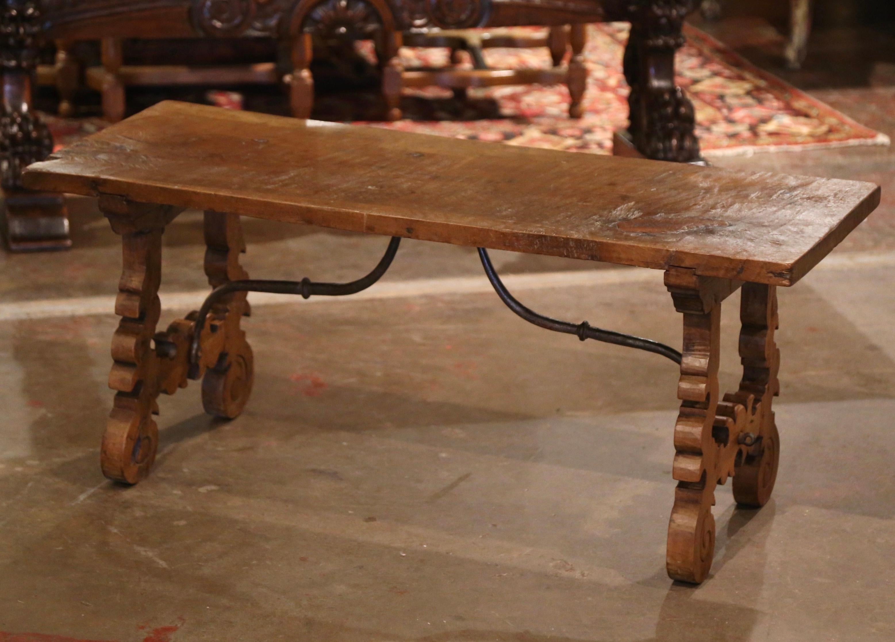Crafted in Spain circa 1780, and built of solid walnut, the rectangular antique trestle coffee table stands on two carved scrolled legs joined together with a forged iron stretcher; the top is built with a single plank of walnut. The long cocktail
