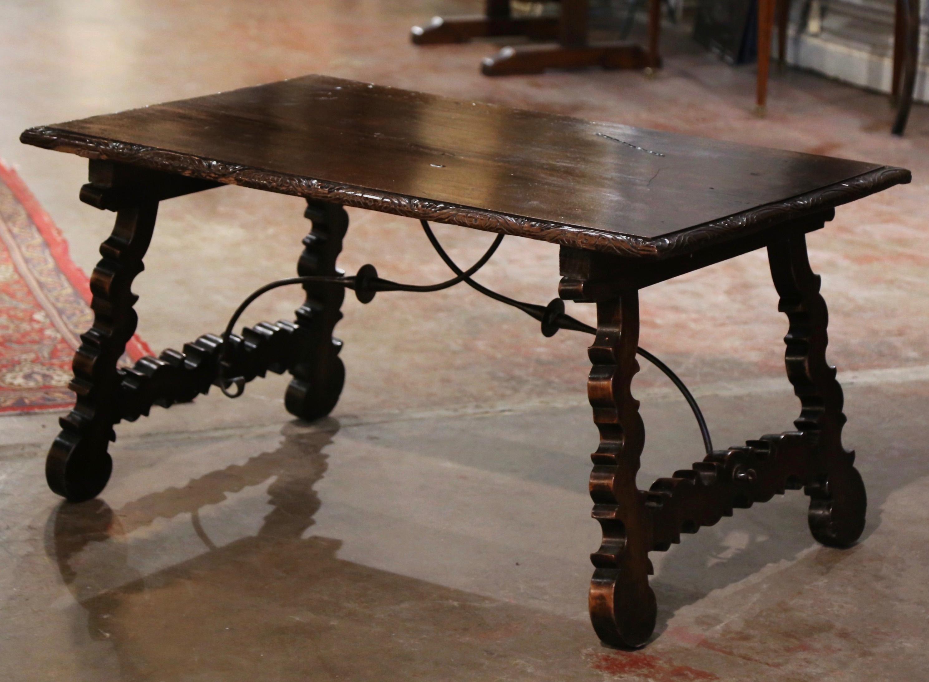 Crafted in Spain circa 1780, and built of solid walnut, the rectangular antique trestle coffee table stands on two carved scrolled legs joined together with a forged iron stretcher; the top is built with two planks of walnut wood and embellished