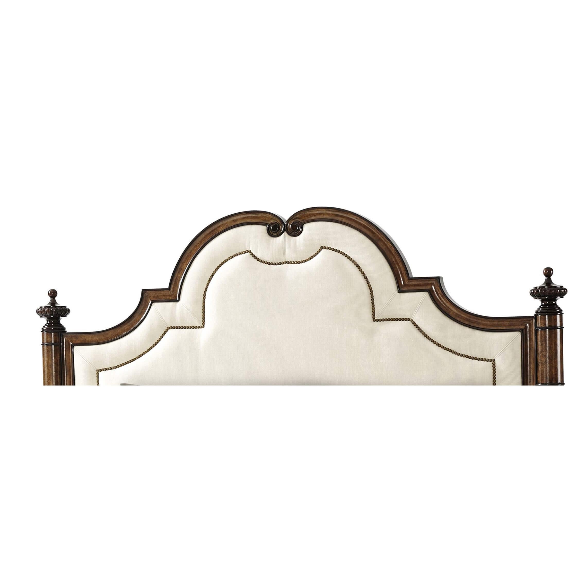 A chestnut burl and flame mahogany veneered bed, the shaped arched headboard with a concave molded frame enclosing a nail head decorated upholstered panel, flanked by ring turned and veneered columns with urn finials, with a similar scroll bracket