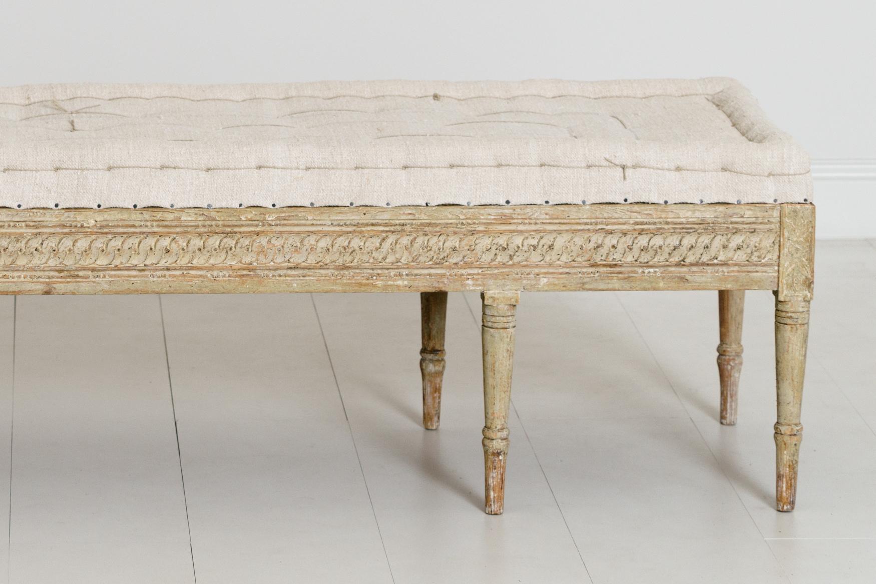 An 18th century Swedish bench from the Gustavian period, newly upholstered in linen with a handstitched tufted seat, circa 1790. There is a carved guilloche pattern on three sides of the seat frame and carved rosettes on the corner posts.
