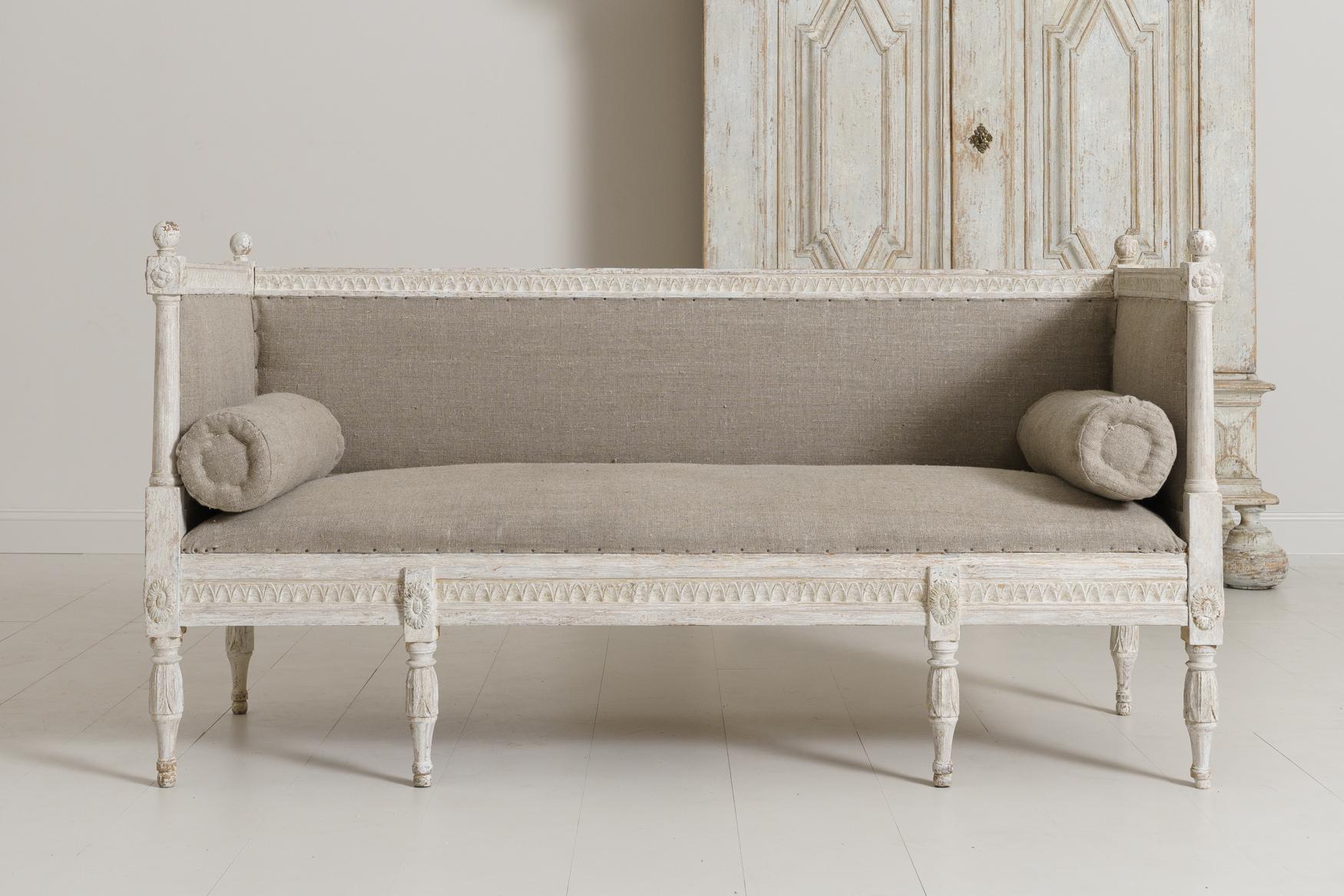 A beautifully carved Swedish sofa bench from the Gustavian period, hand-scraped to reveal the original paint surface and newly upholstered in linen with side bolster pillows, circa 1800. The back and seat frame feature carved lamb's tongue detail