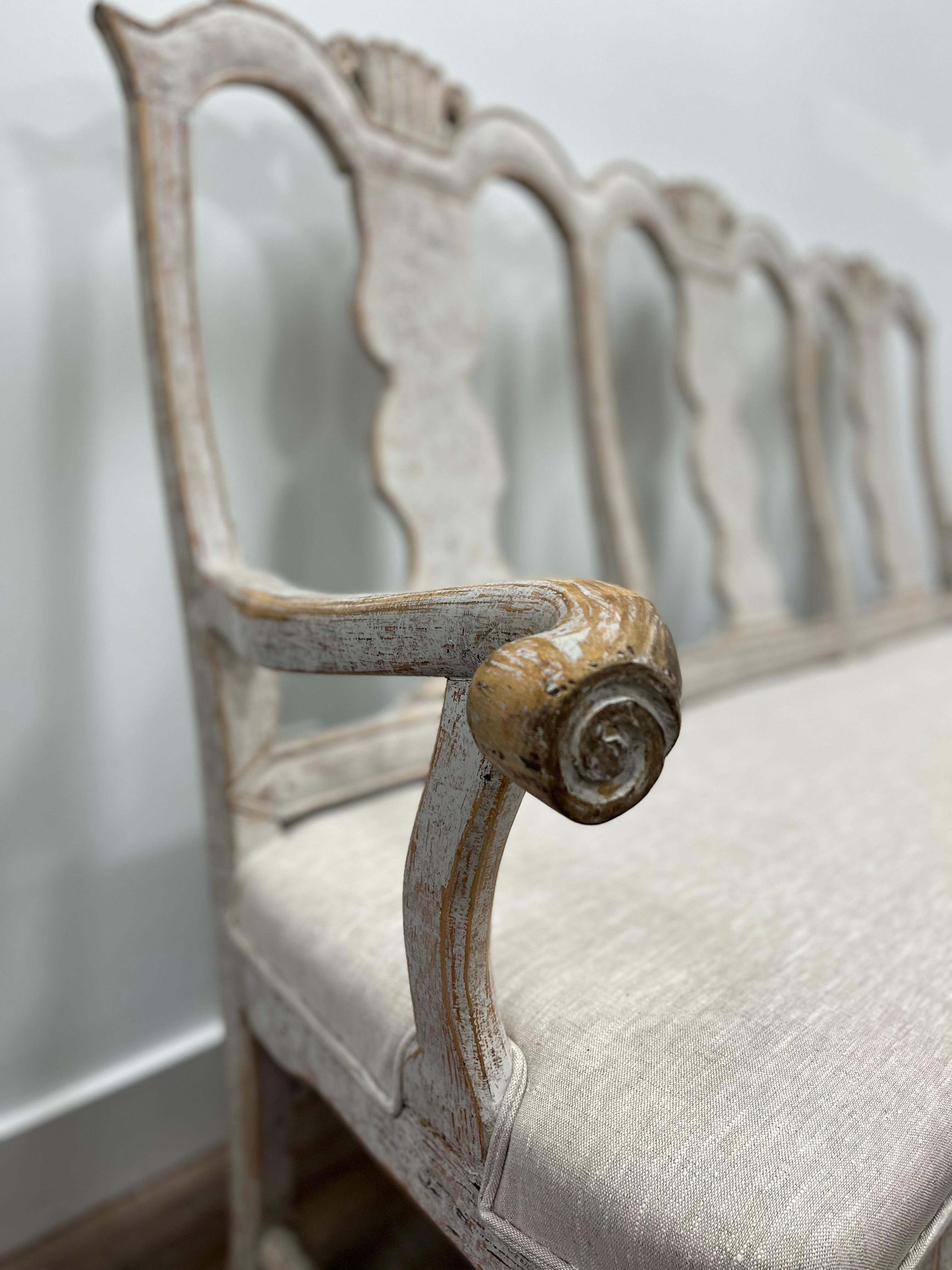 A Swedish Baroque sofa with outward bending arms and hand-turned ends. The arched back crowns have simple scroll detailing. Notches line the contours of the spines and back frame. A hand-carved swag squirt front with traces of vertical detailing.