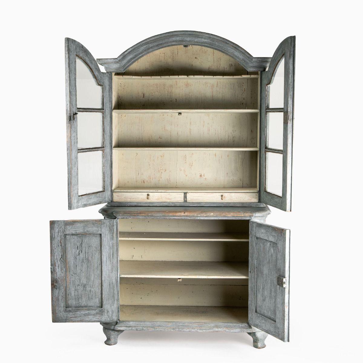 Vitrina rococó fabricada en pino con un bonito acabado pintado en gris azulado.

El armario se compone de dos partes:
La parte superior presenta un elegante arco con una cornisa perfilada y un par de puertas de paneles acristalados que siguen el