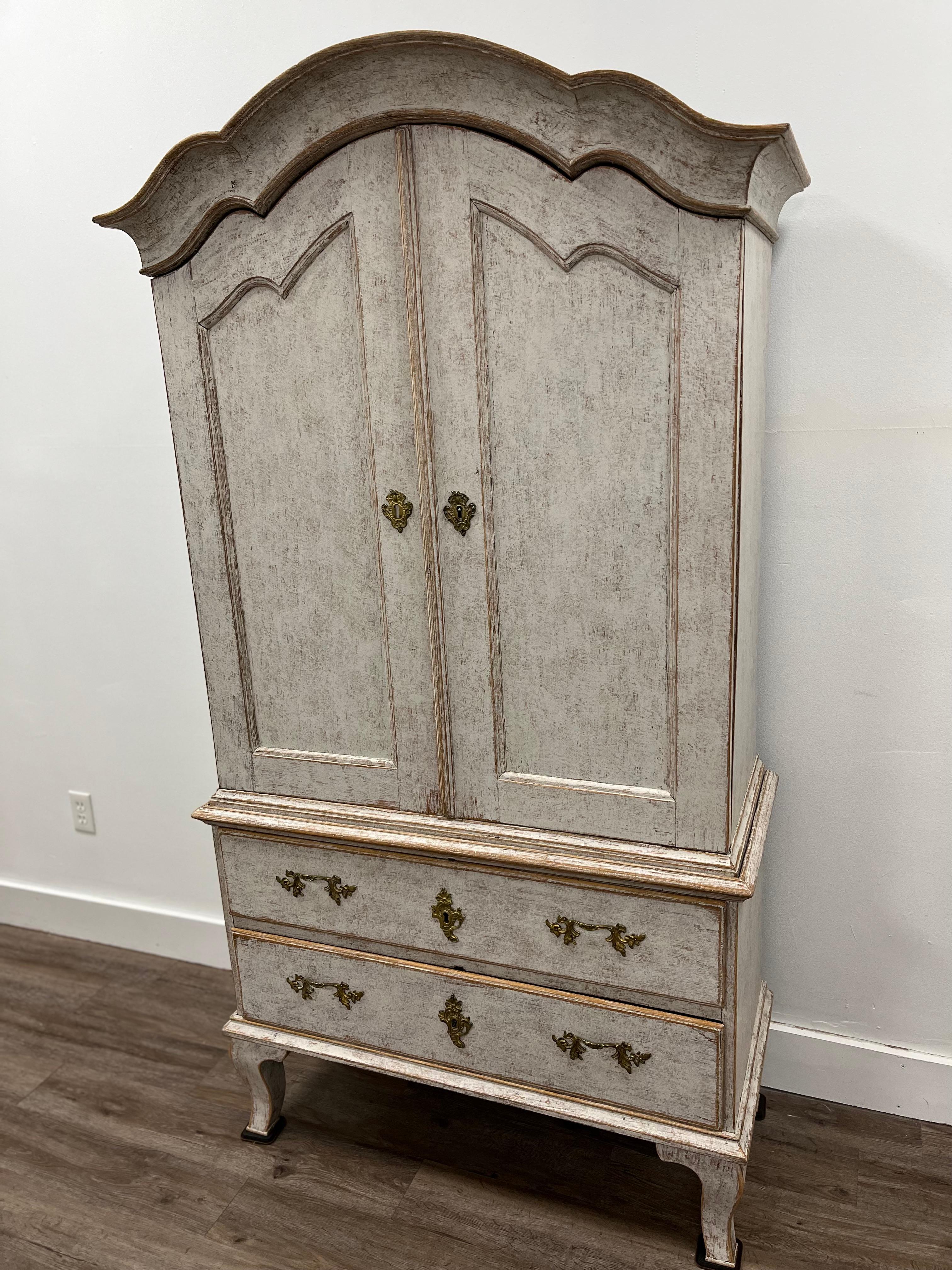 A demure Swedish Rococo cabinet with an impressive overhanging arched pediment cornice bonnet top. Upper section has four shelves and three small drawers with brass pulls. Door fronts have detailed recessed wood carvings. Lower section has two