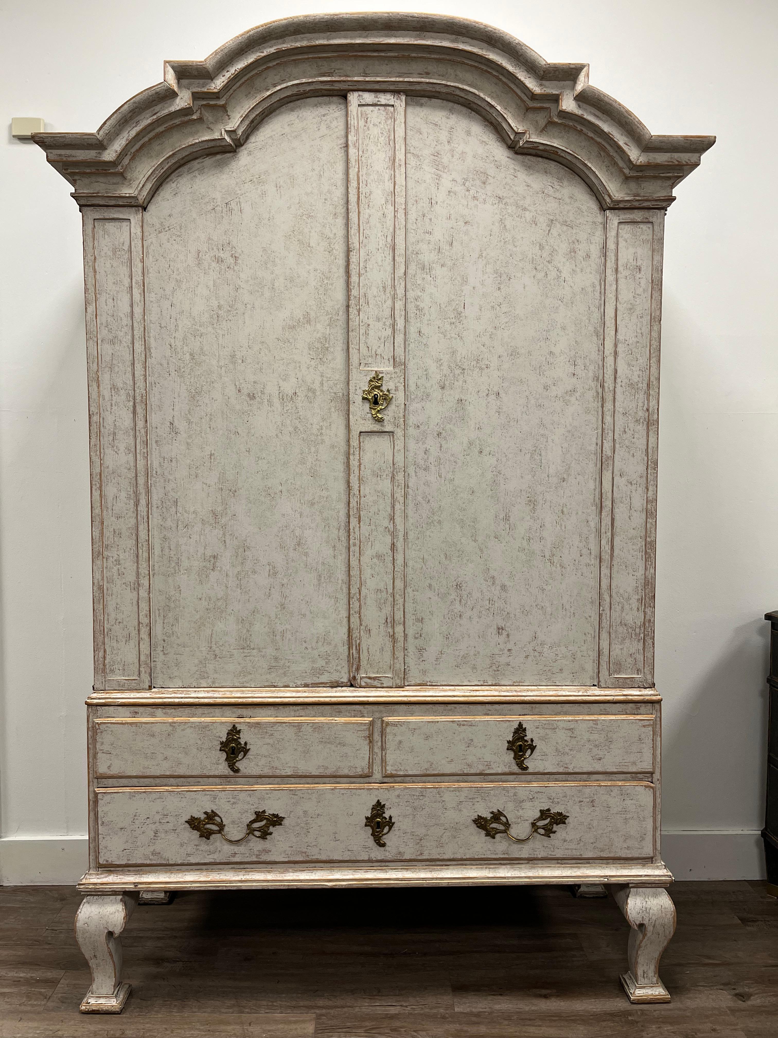 A Swedish Rococo cabinet. The bonnet top consists of an impressive overhanging arched pediment cornice. Upper section has three  shelves and three small drawers (with teardrop brass pulls). Lower section has two small drawers atop one large drawer.