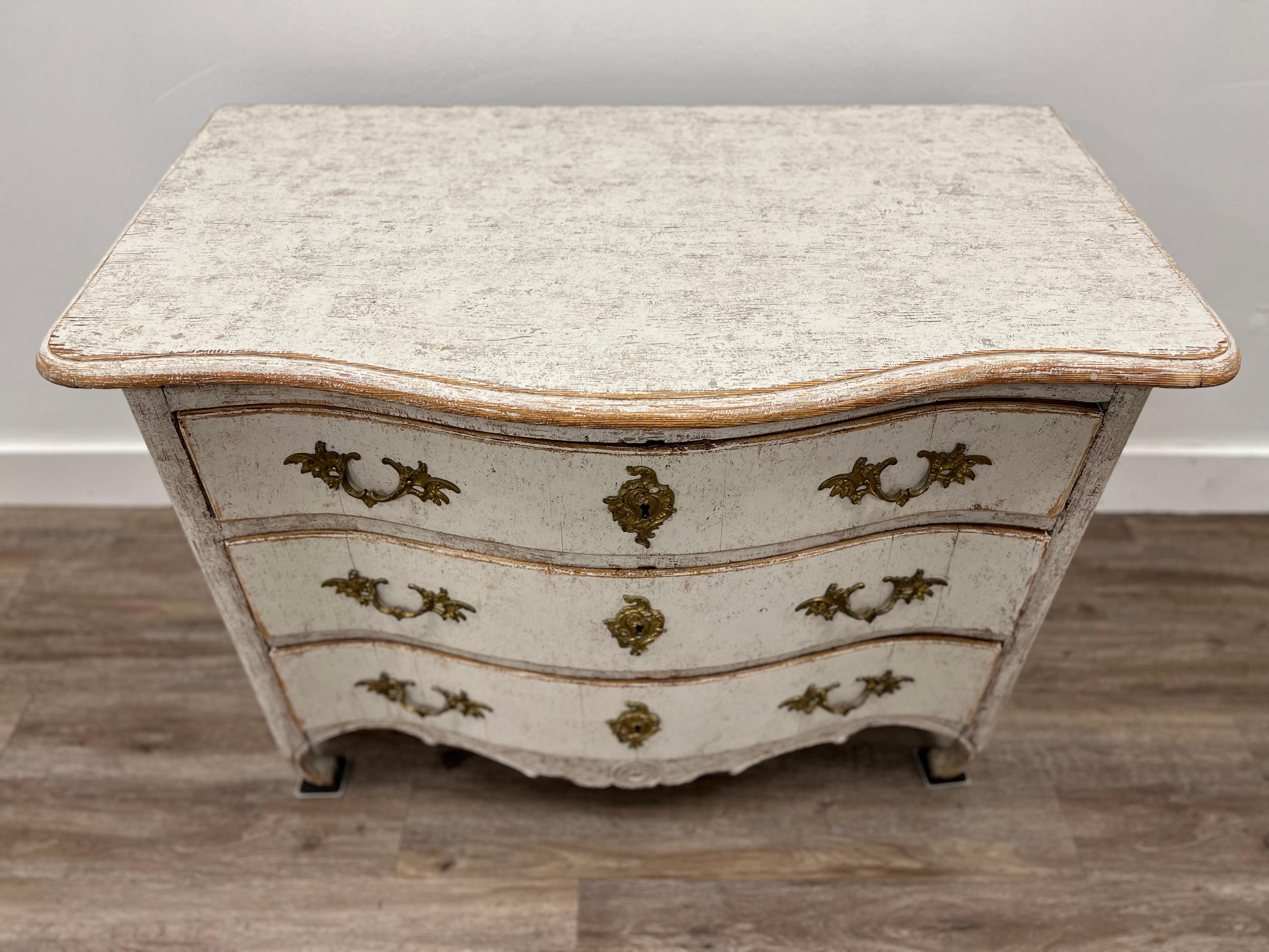 A Swedish Rococo commode with a slide-out tray over three bow front drawers. Decoratively carved apron. Repainted in soft grey with original locks and old (but not original) brass hardware. Original freight goods label on the back.