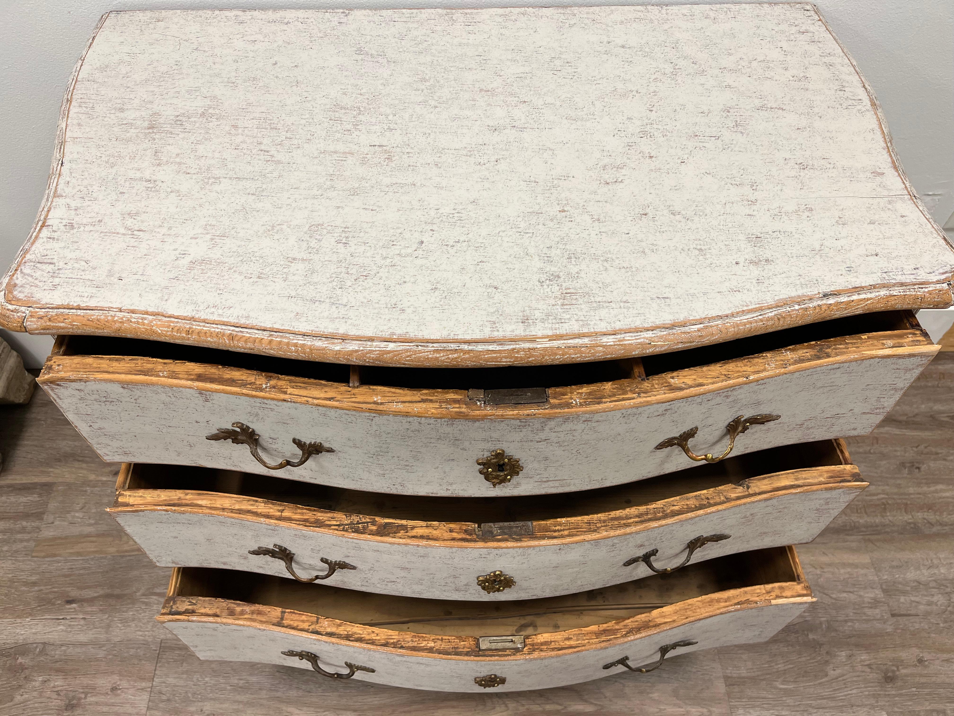 A Rococo commode with original brass pulls, hardware and locks on three bow front drawers. The top drawer has three compartments. Commode top front and sides are contoured. Apron and feet are shaped. Tastefully repainted in light grey.