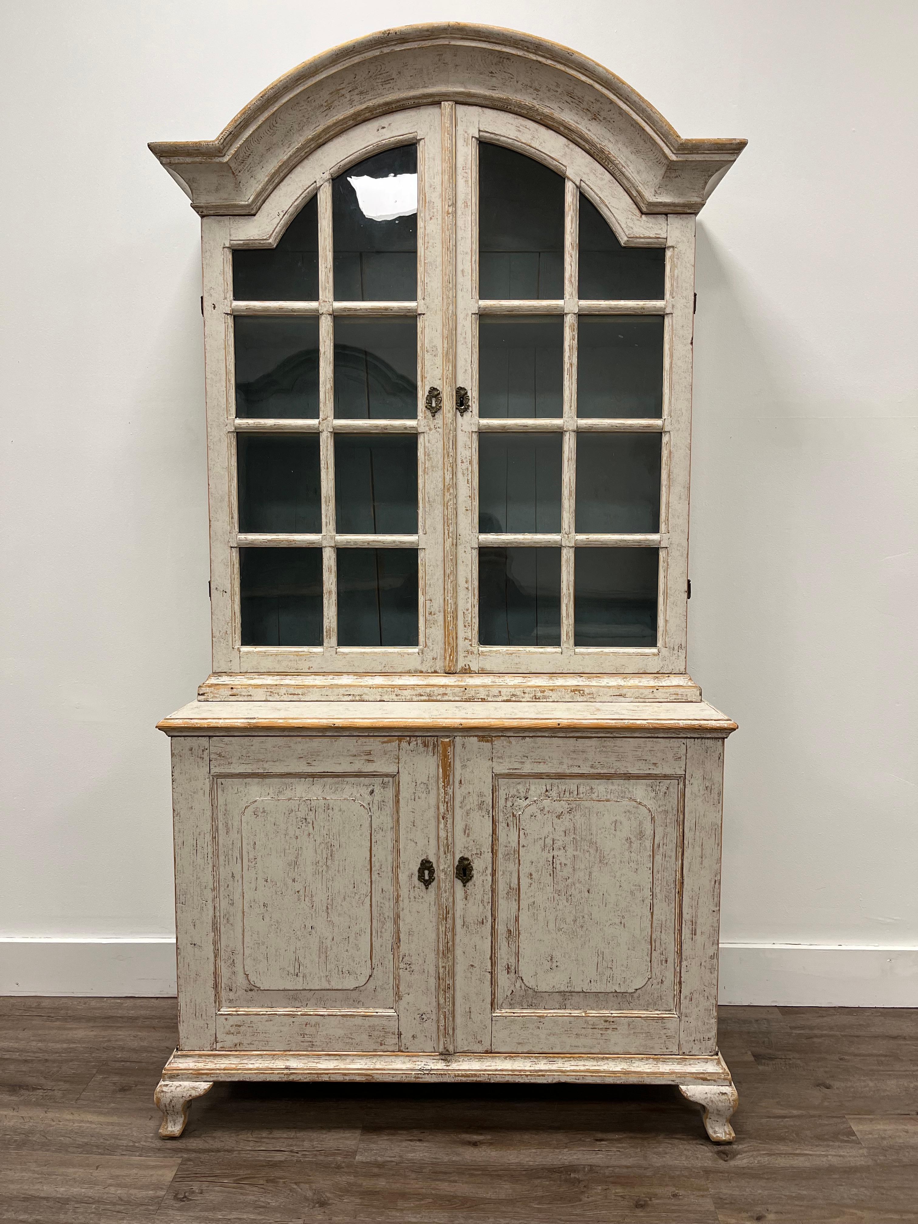 A beautiful Rococo vitrine with original brass hardware and locks. The upper cabinet has four shelves under a bonnet top with glass doors. The lower cabinet has two shelves behind raised panel doors. Sits atop cabriolet feet. Tastefully repainted
