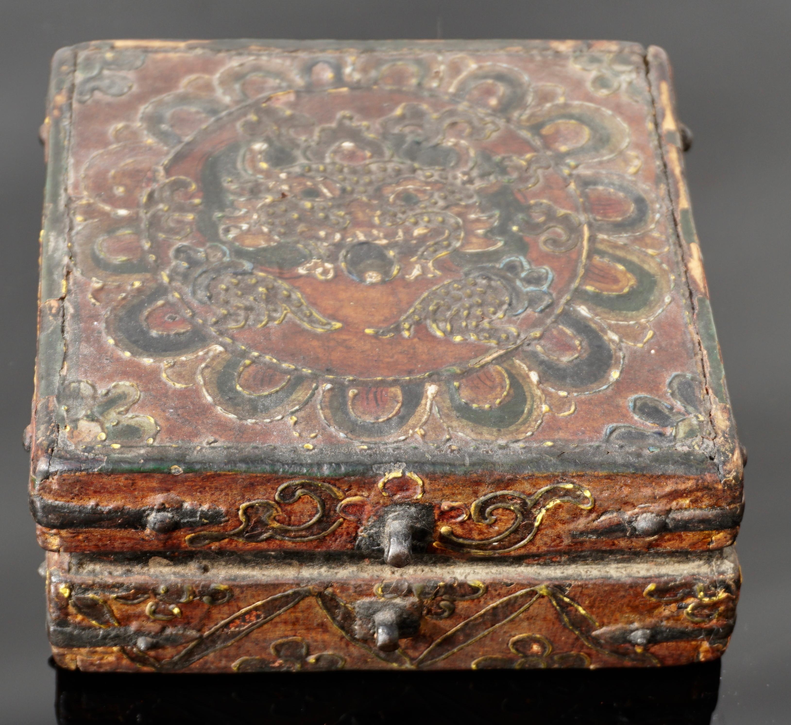 A small and very charming Tibetan covered box constructed with metal straps and painted with thick polychrome paint. The decorative scroll designs culminate into a Buddha or wrathful deity with extended hands encircled by a Lotus flower in ox blood