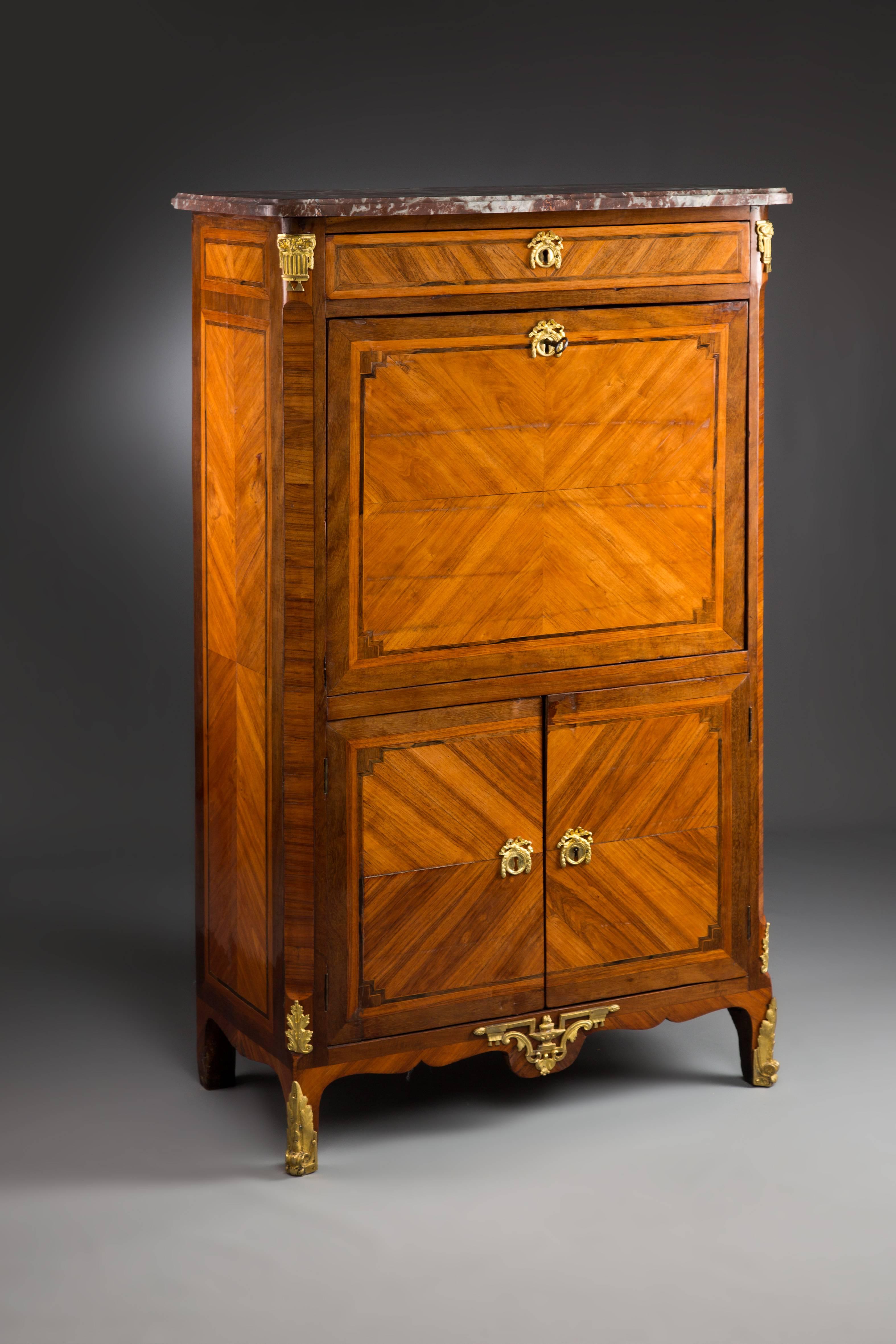 Rose veneer on oak. High-rectangular body with finely veneered wood on four feet. Foldable writing board (leather cover missing), a drawer, lower wing door. Gilded bronze mountings. Top cover with a gray brown marble top. Signed.


(L-128).