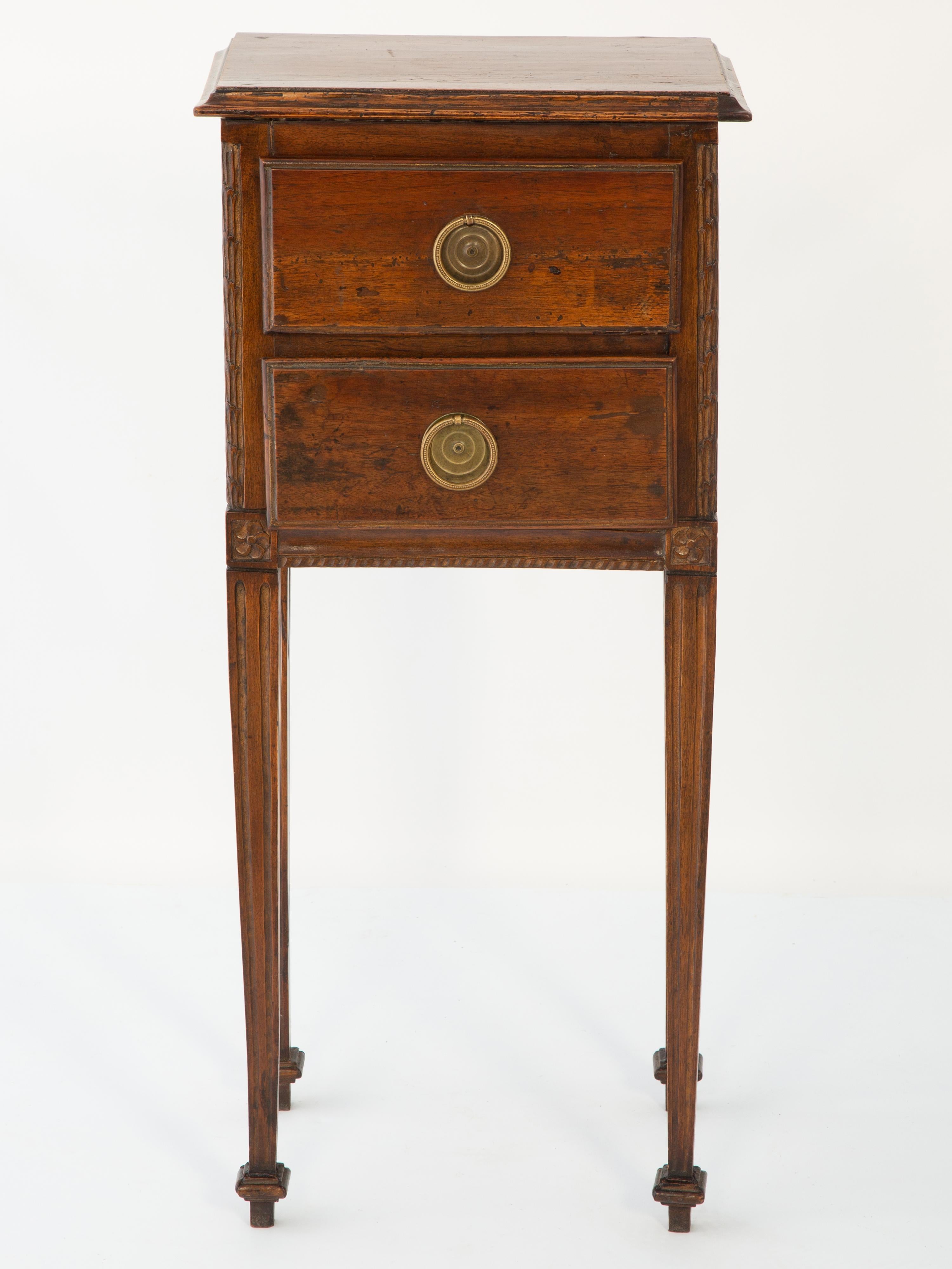 Petite bedside nightstand or side table commode of walnut. Its rectangular molded top, over two stacked drawers with round brass pulls. The drawers are flanked by stiles carved with husking, over gadrooned apron. Raised on four, tapering,