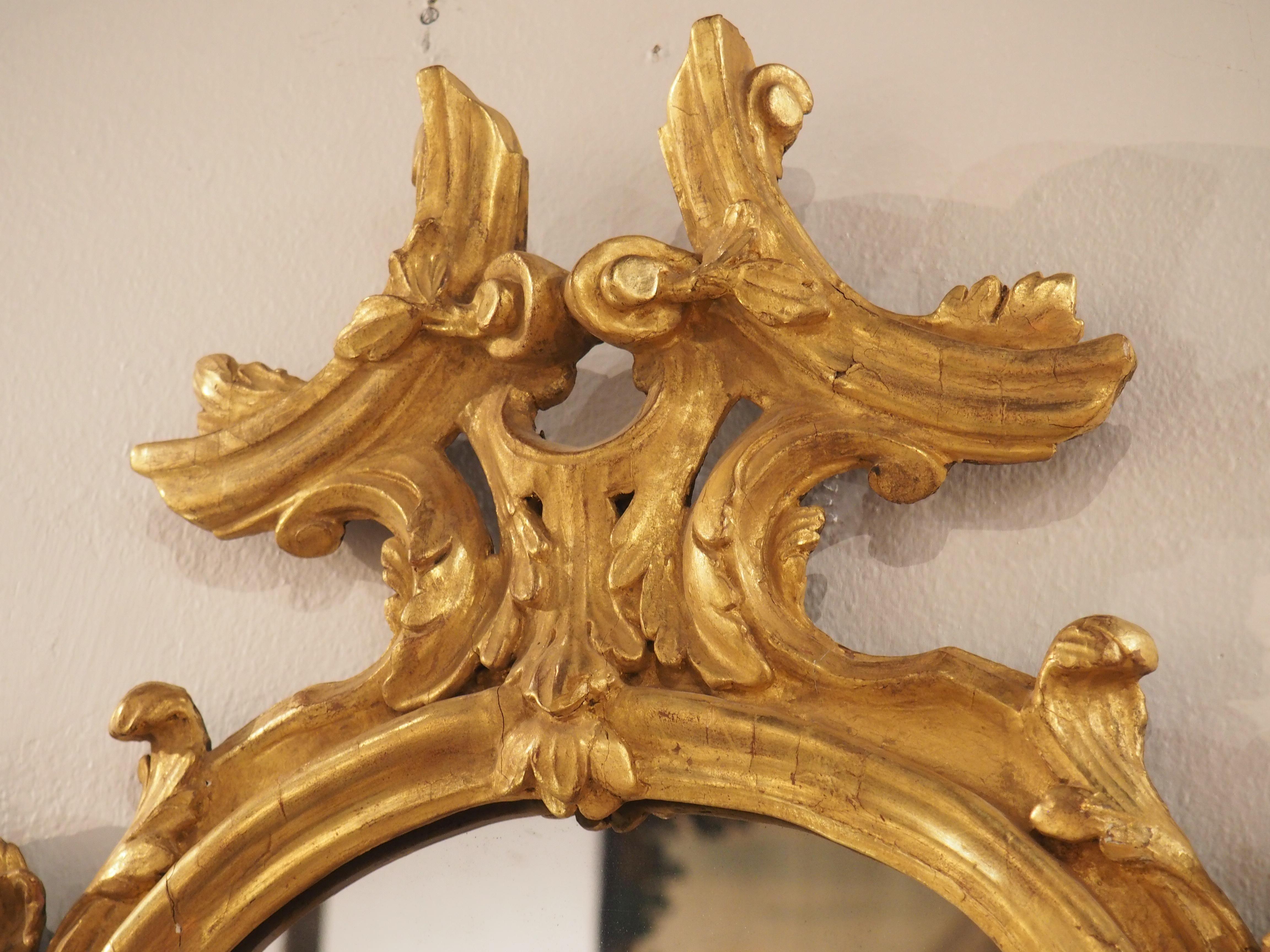 Hand-carved in the Veneto (or Venetia) region of North Italy, this gorgeous, giltwood mirrored wall sconce is from the mid-1700s. Two arms slot into a hole in the center of a curled leaf at the bottom of the mirror. The curvaceous arms terminate in