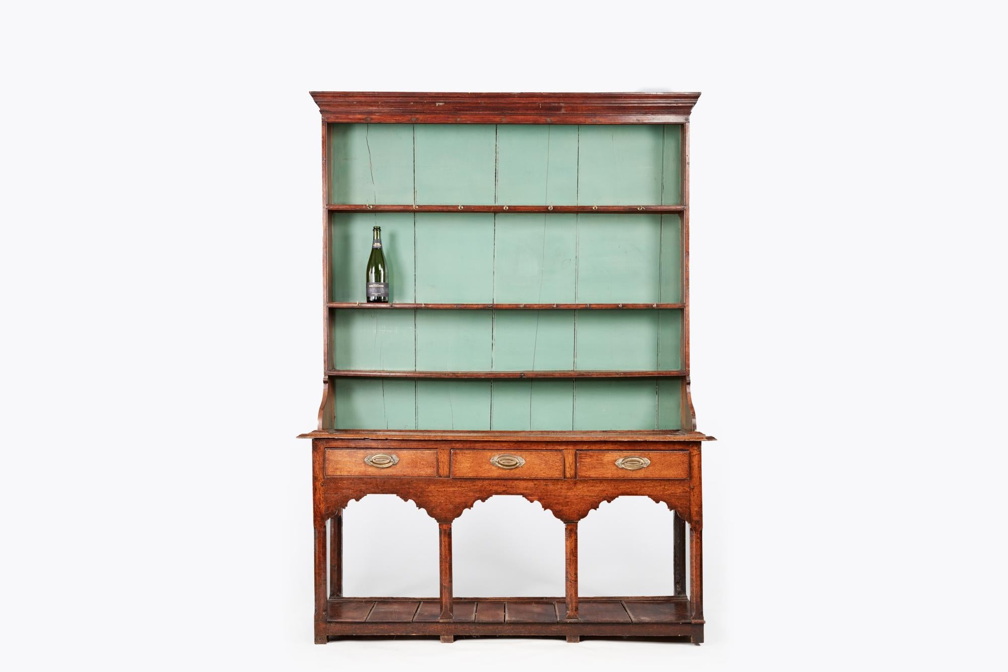 18th century Welsh potboard dresser of mixed oak, ash and painted pine with a pot board base. It features three central drawers with brass handles and a carved apron to the front sitting above three arches with chamfered supports. The upper section