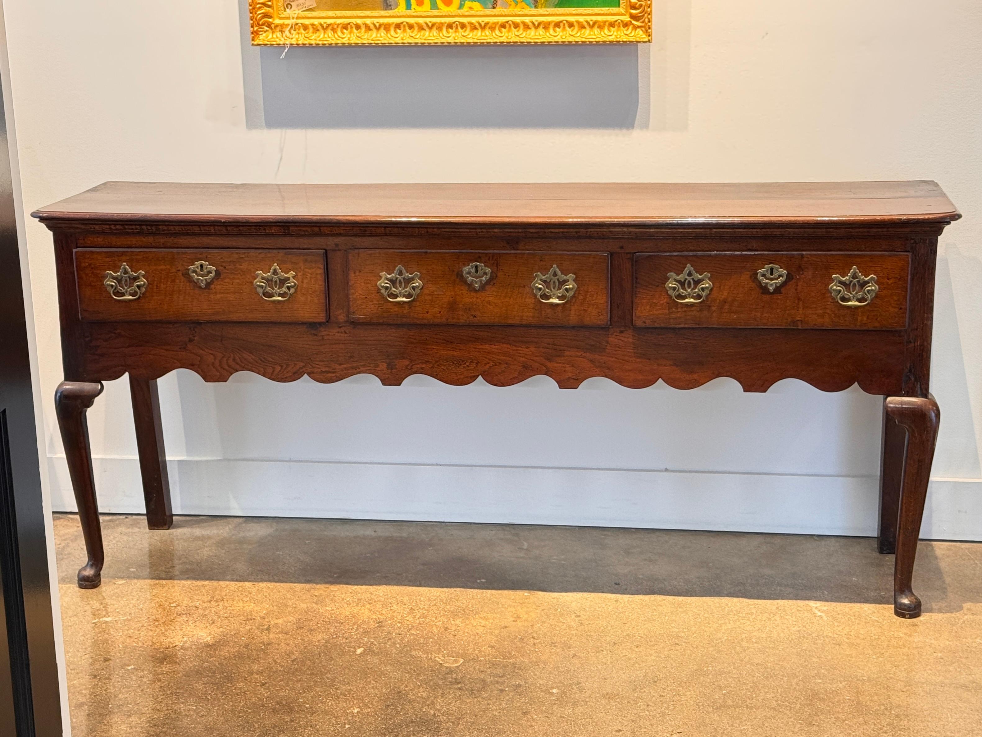 A 19th century Welsh oak dresser base with four drawers and a boldly scalloped apron. The rich patina and brass hardware highlight the natural grain and age of the wood. Cabriole front legs and shaped ends contribute to the piece’s traditional