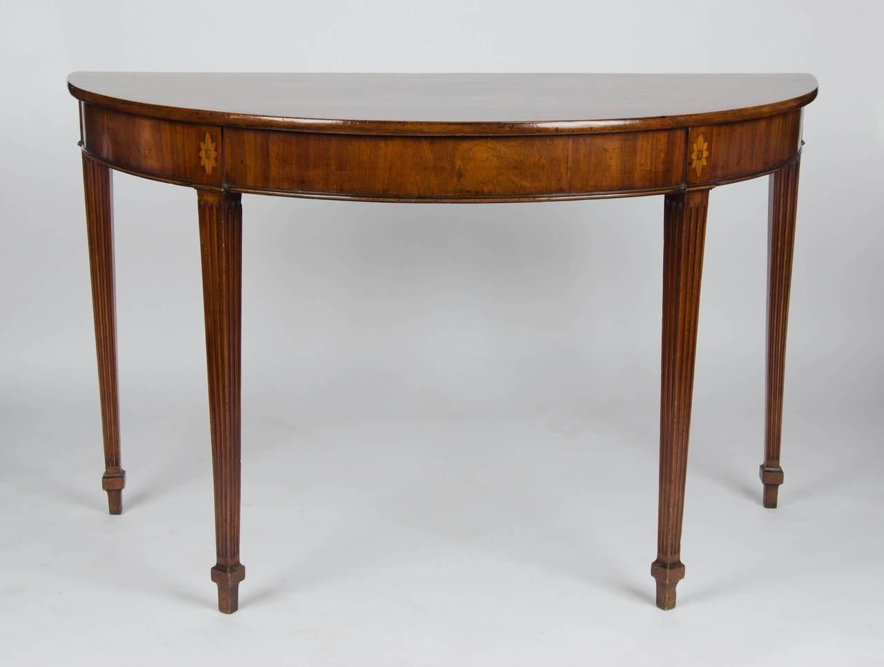 A wonderful late 18th century, mahogany demilune side or serving table. 
The well figured top above a frieze with floral paterae inlay upon fluted tapering lags with block and spade feet.