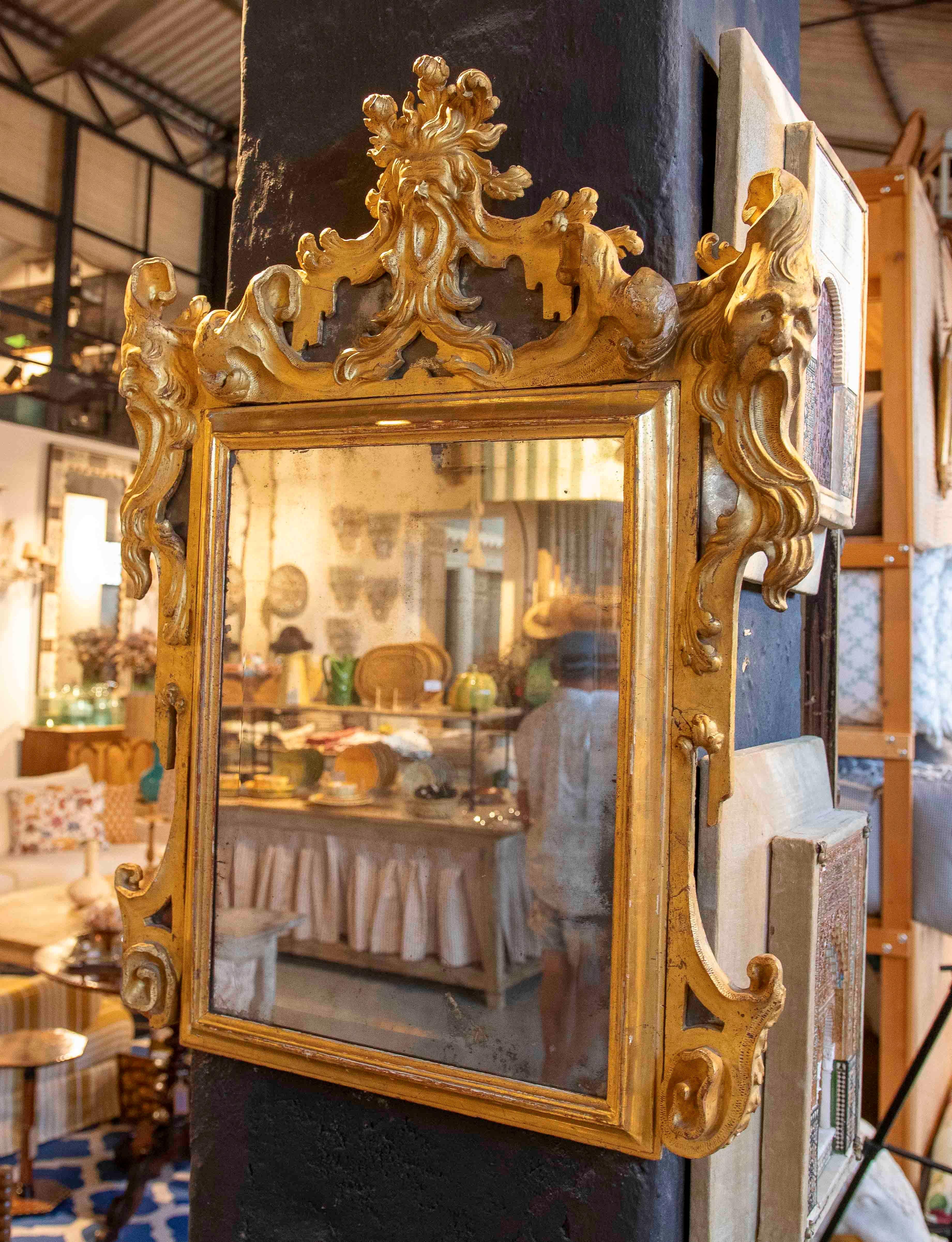 An elegant Rococo-style mirror, crafted from carved wood and covered with gold leaf. The frame features elaborate scrollwork and stylized female figures on the sides, crowned by a central foliage and cartouche motif characteristic of the late