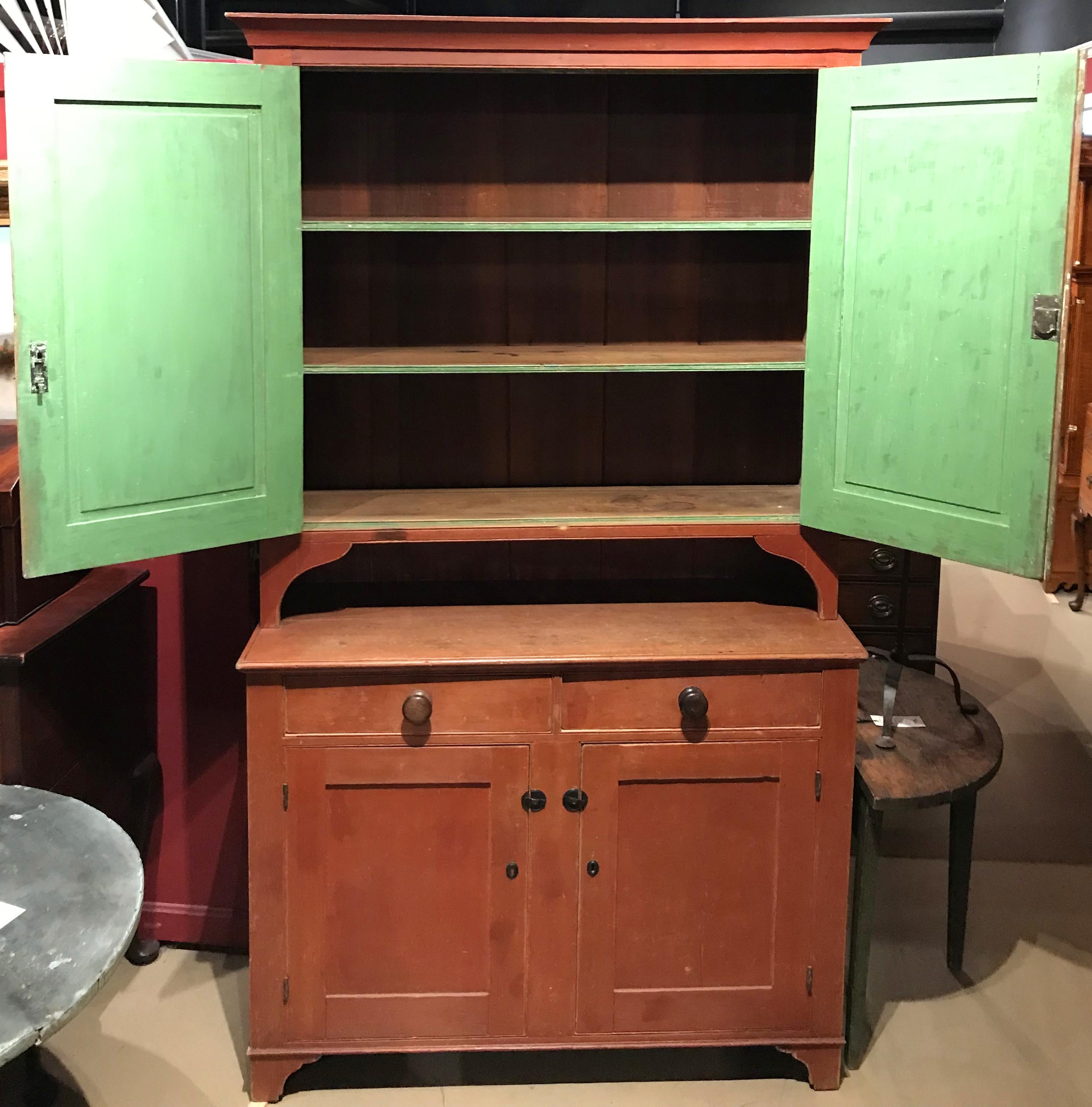 A wonderful two part stepback cupboard in old red, the upper case with molded cornice surmounting two paneled doors, opening to reveal shelves with an old green painted interior. The upper cabinet is integrated with the counter/top board of the