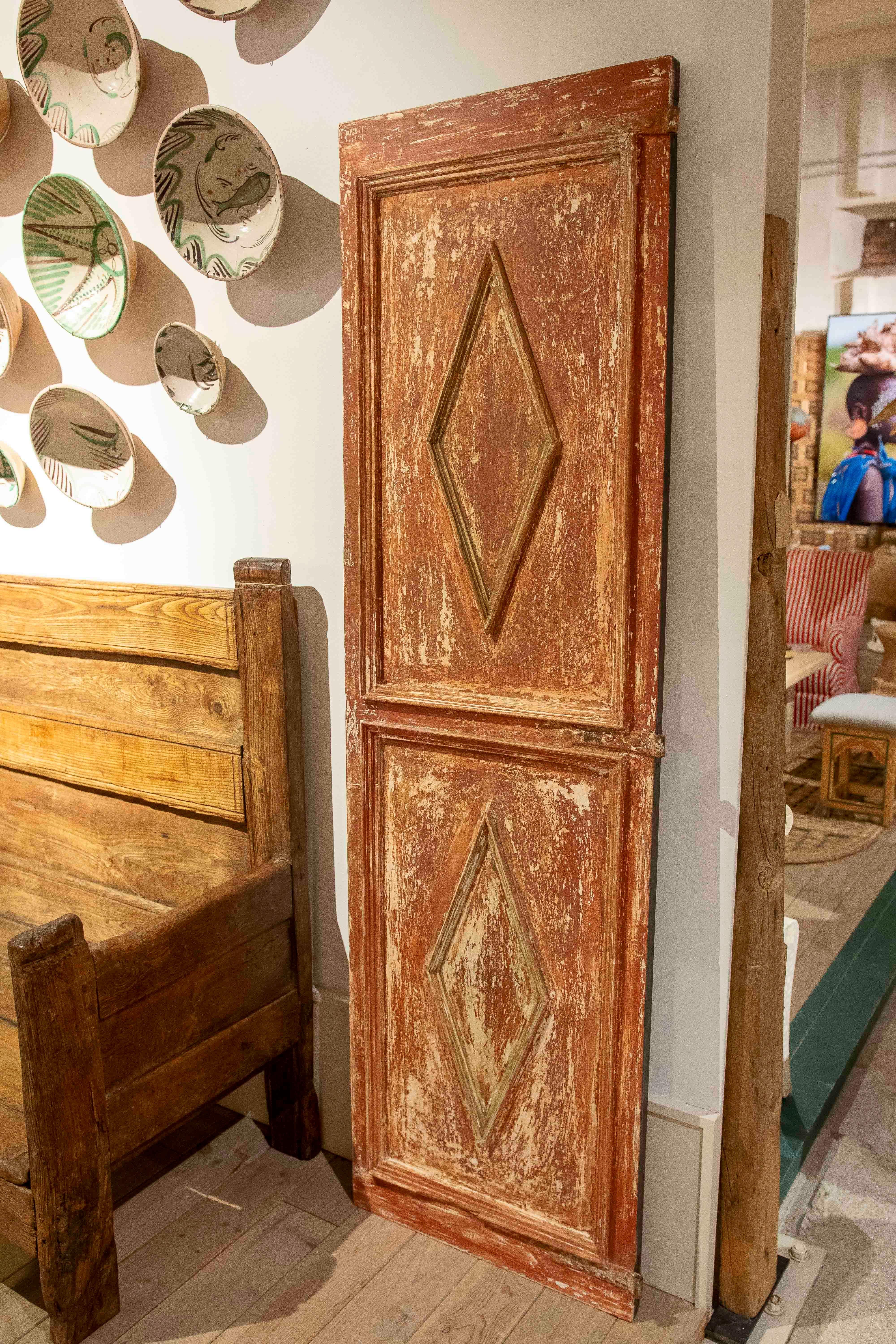 A set of two antique solid wood doors, each composed of two panels with a molding and a central carved diamond-shaped decoration. The surface has an original patina with natural wear, in reddish and ochre tones, lending authenticity and character to