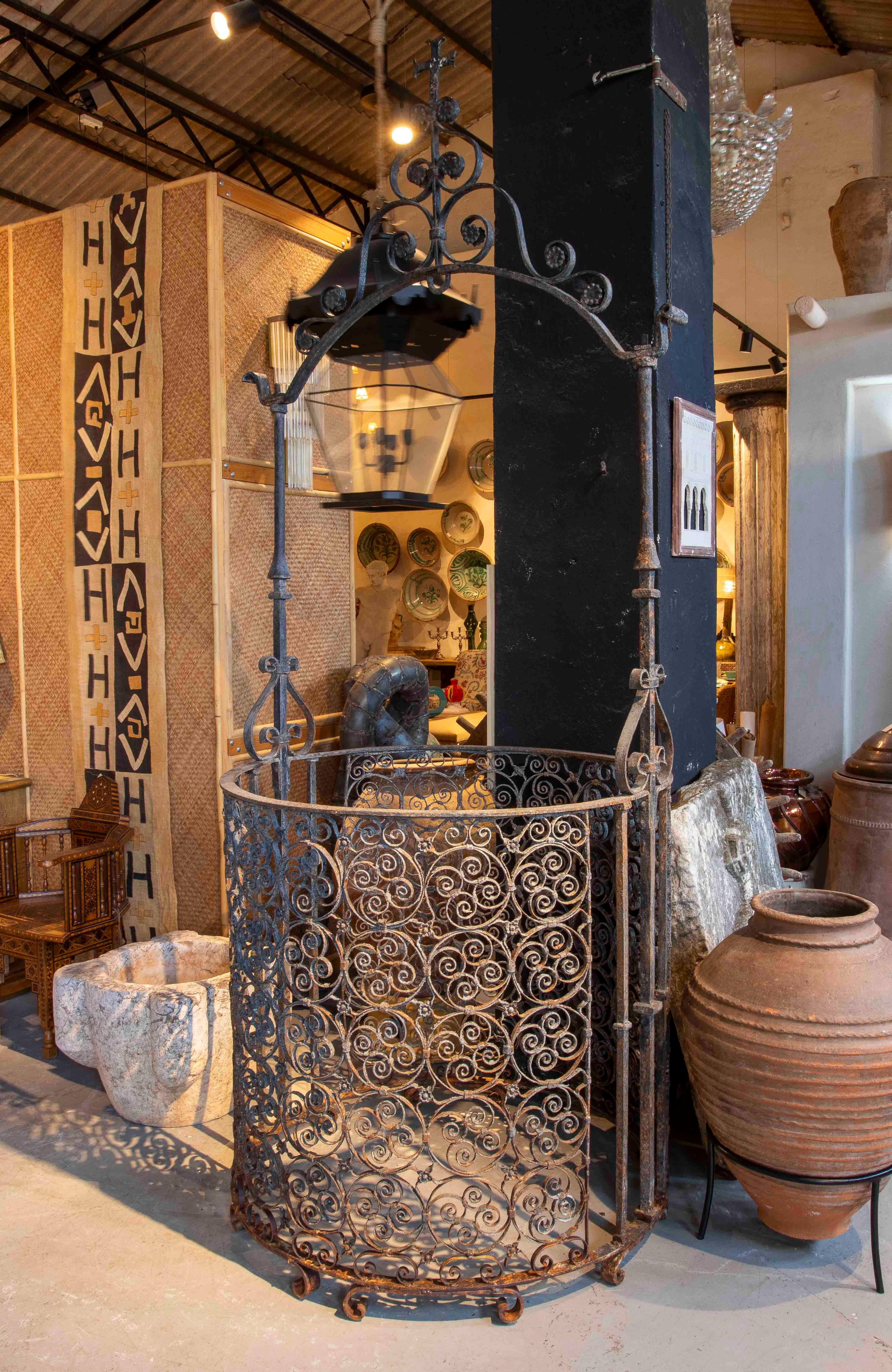 A stunning decorative wrought iron well with a cylindrical body featuring intricate scrollwork motifs that give it a romantic, handcrafted feel. The piece is crowned by an ornately detailed arch and an elegant hanging lantern, making it an ideal