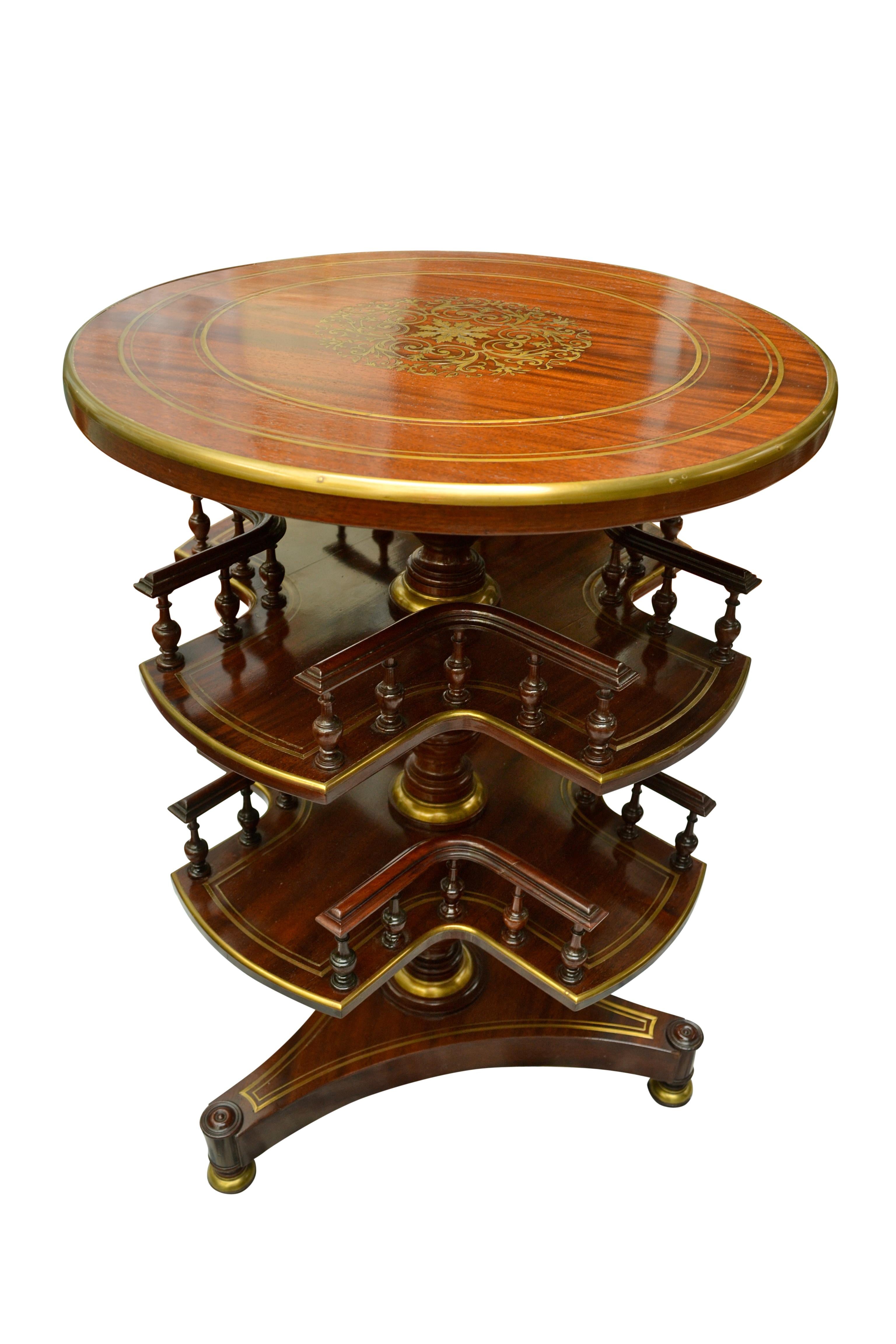 A circular mahogany and inlaid brass library table, with two tiers of revolving book/CD/liqour bottle and glasses shelves below the fixed top, all supported on a circular turned column joining a tri-form base. The top has a central brass inlaid
