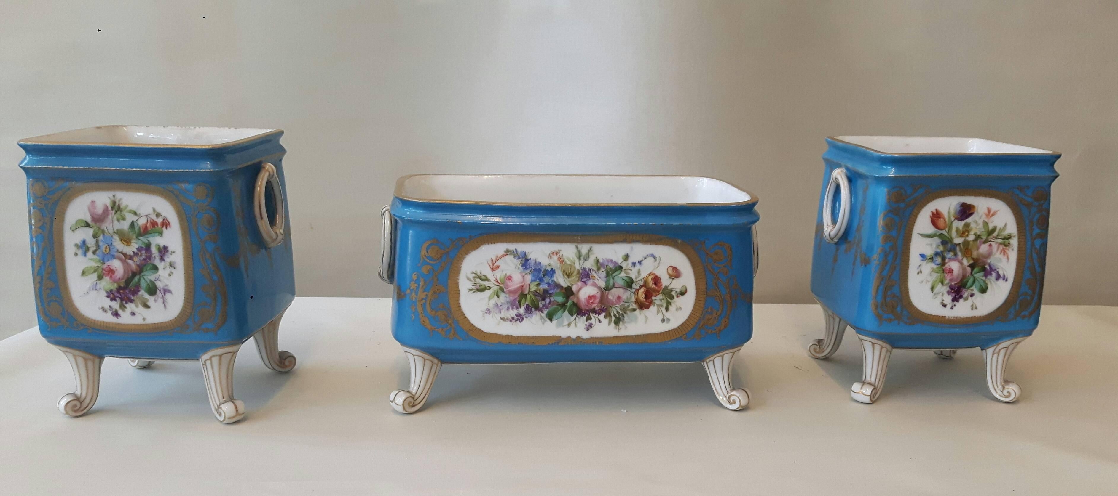 An unusual trio of jardinière and cache-pots, in the Sevres style, hand-painted with love scenes after Watteau on one side whilst the other side is decorated with flower panels. Each piece rests upon scrolled feet, and has tromp l'oeil handles on