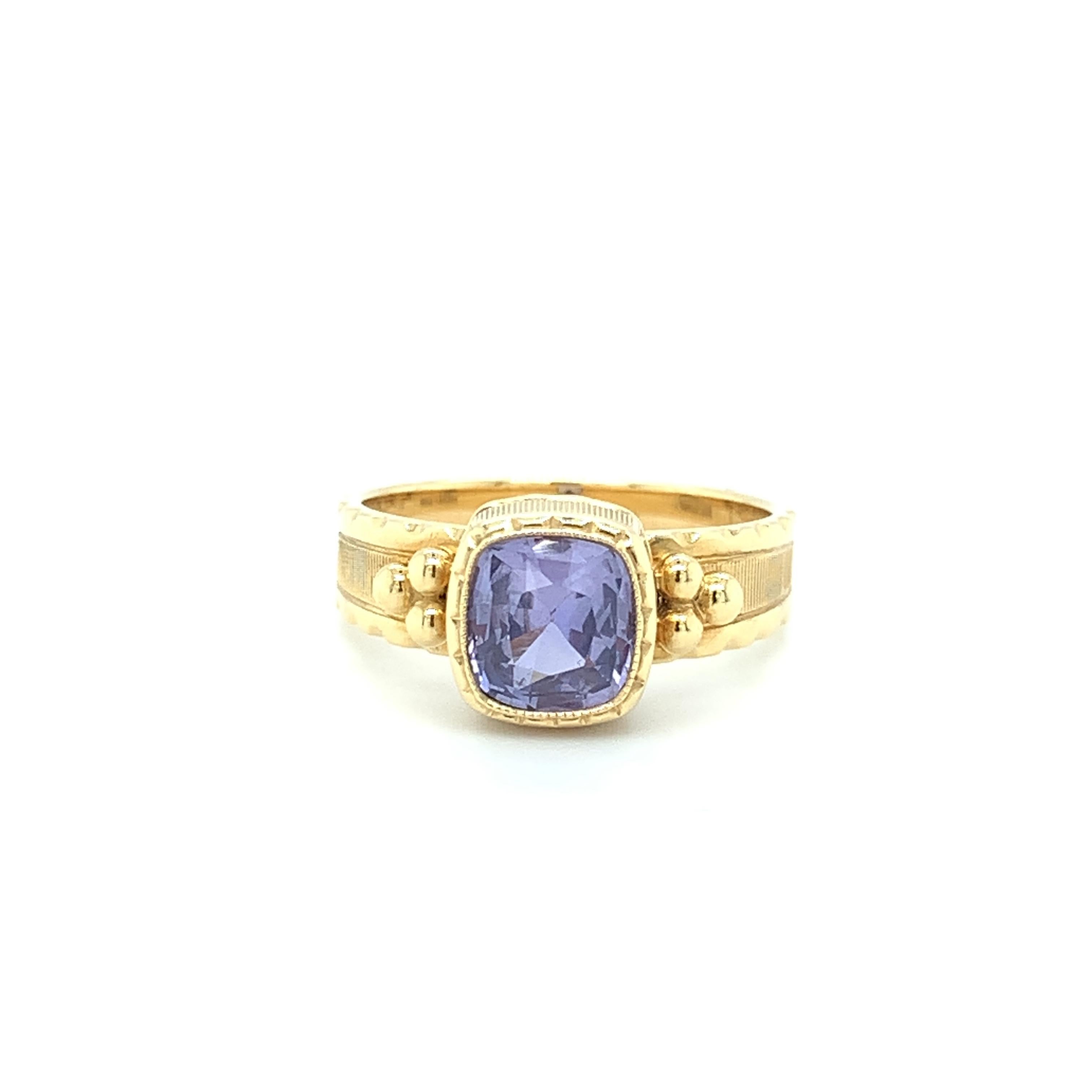 This pretty ring features a lively 1.90 carat fancy color violet sapphire that has been set in a handmade 18k yellow gold bezel. The sapphire is cushion shaped and it is a beautiful shade of bright, bluish purple. It is accented on both sides with