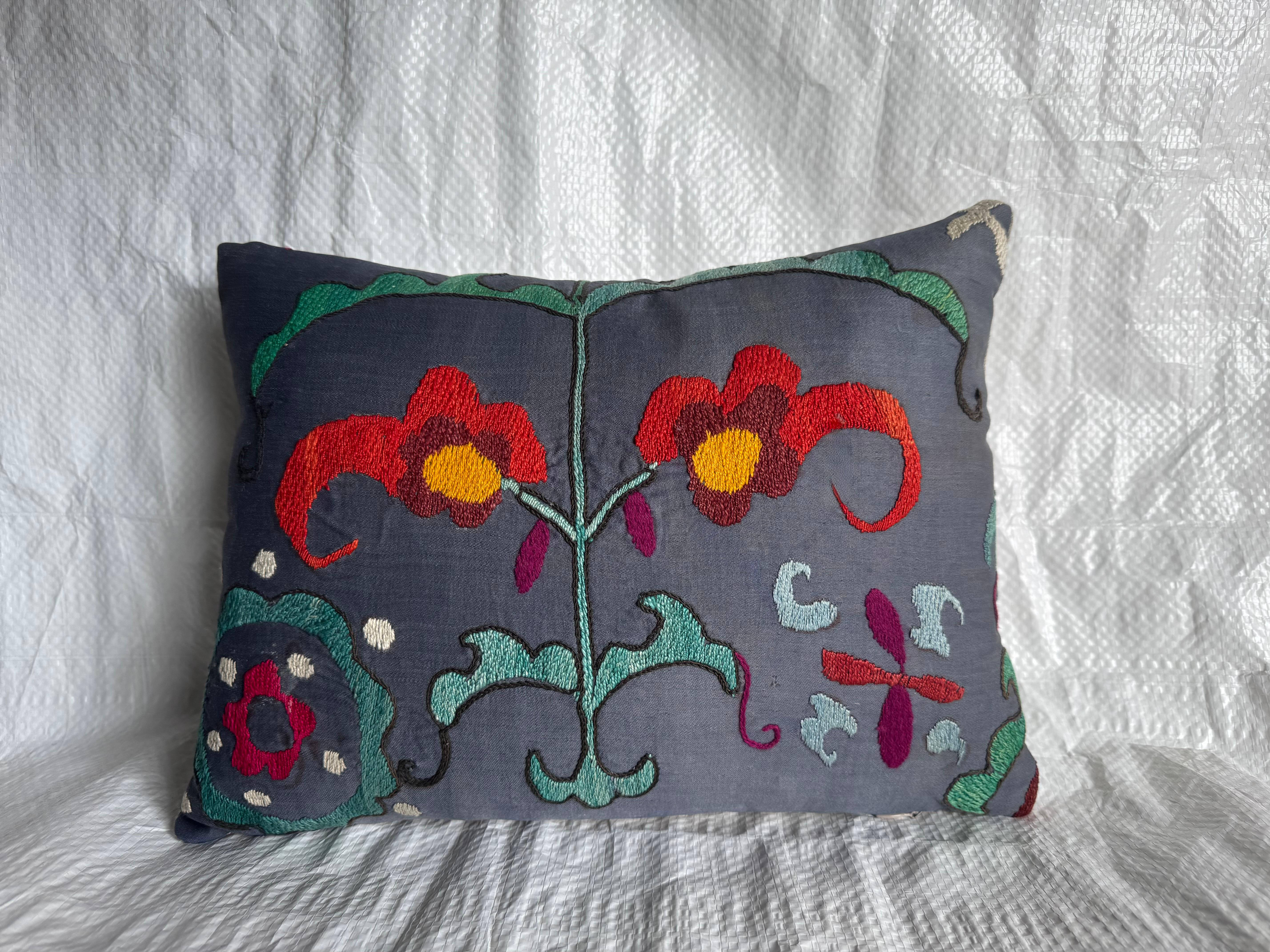 A striking vintage Suzani pillow showcasing bold Central Asian embroidery on a soft charcoal base. The hand-stitched design features vivid red and gold floral motifs with accents of turquoise and plum, representing traditional Suzani symbolism of