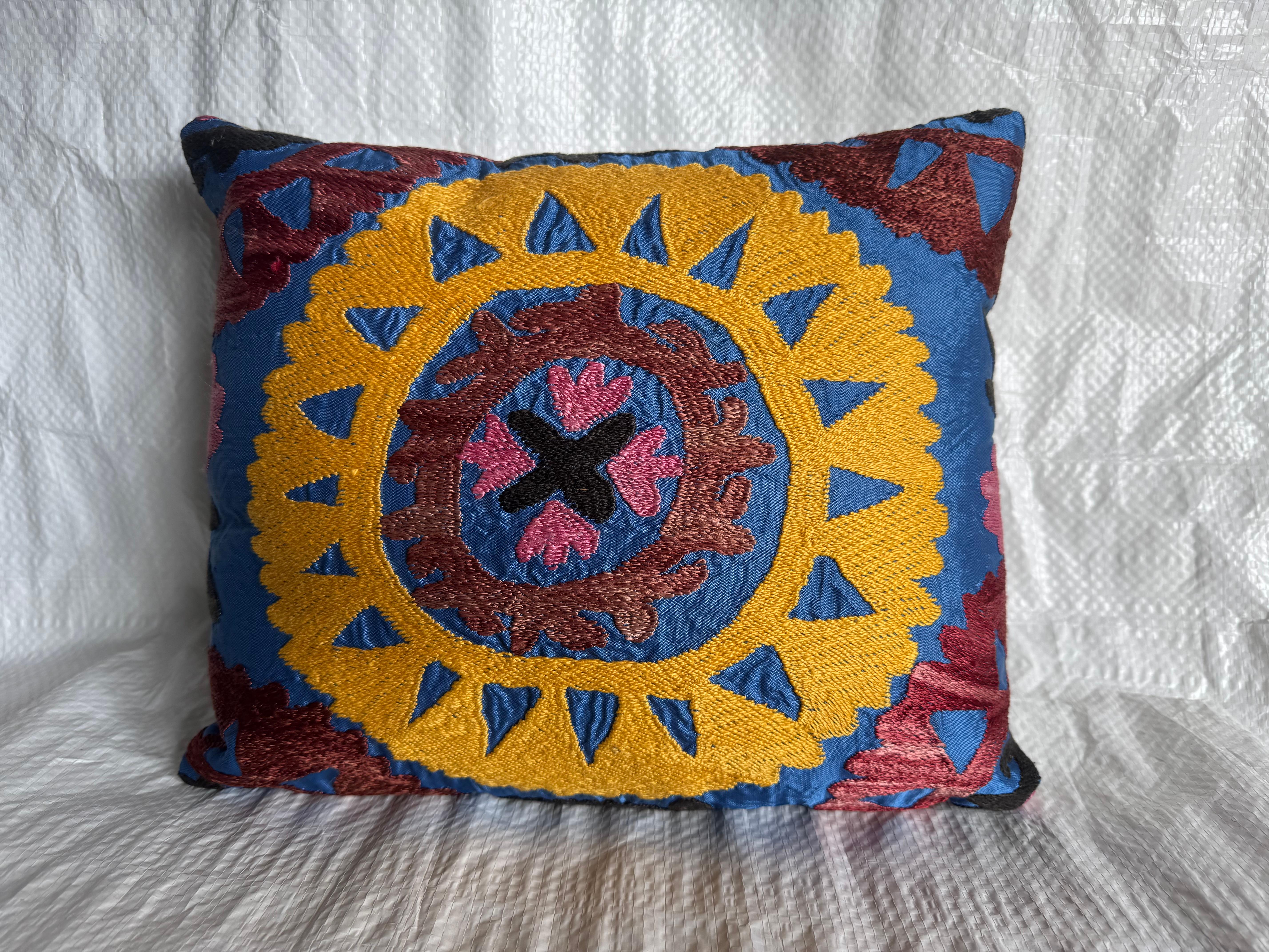Un superbe coussin Suzani antique des années 1900 présentant de vibrantes broderies à la main en moutarde dorée, bordeaux, rose et noir sur un riche fond bleu. Le médaillon circulaire complexe est un motif classique des textiles d'Asie centrale,