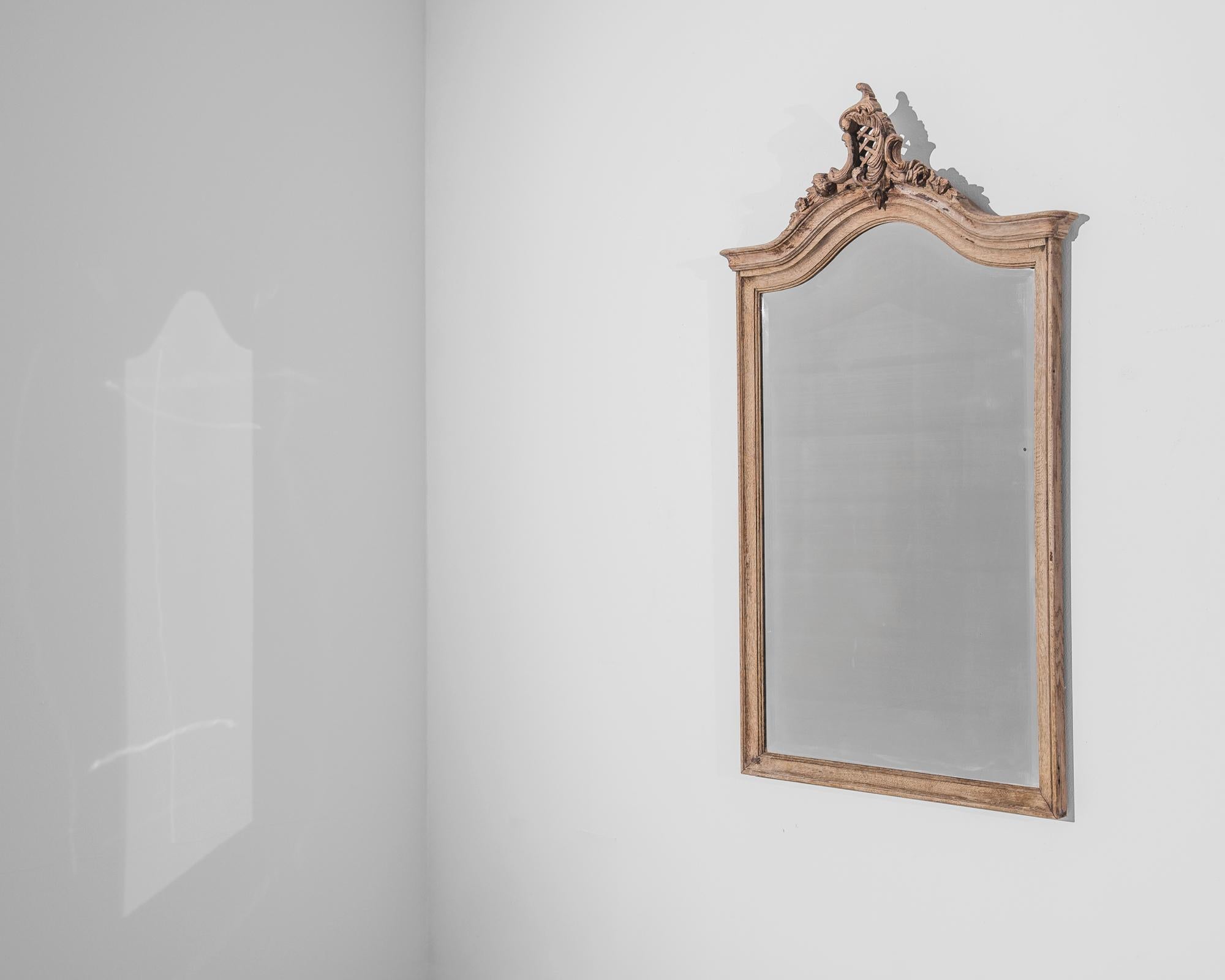 A wall mirror from 1920s France with a carved oak frame. The rectangular frame arches at the top, crowned with a stylized decoration subtly mimicking natural forms. The oak frame has a natural finish and a warm russet tone, accentuating the