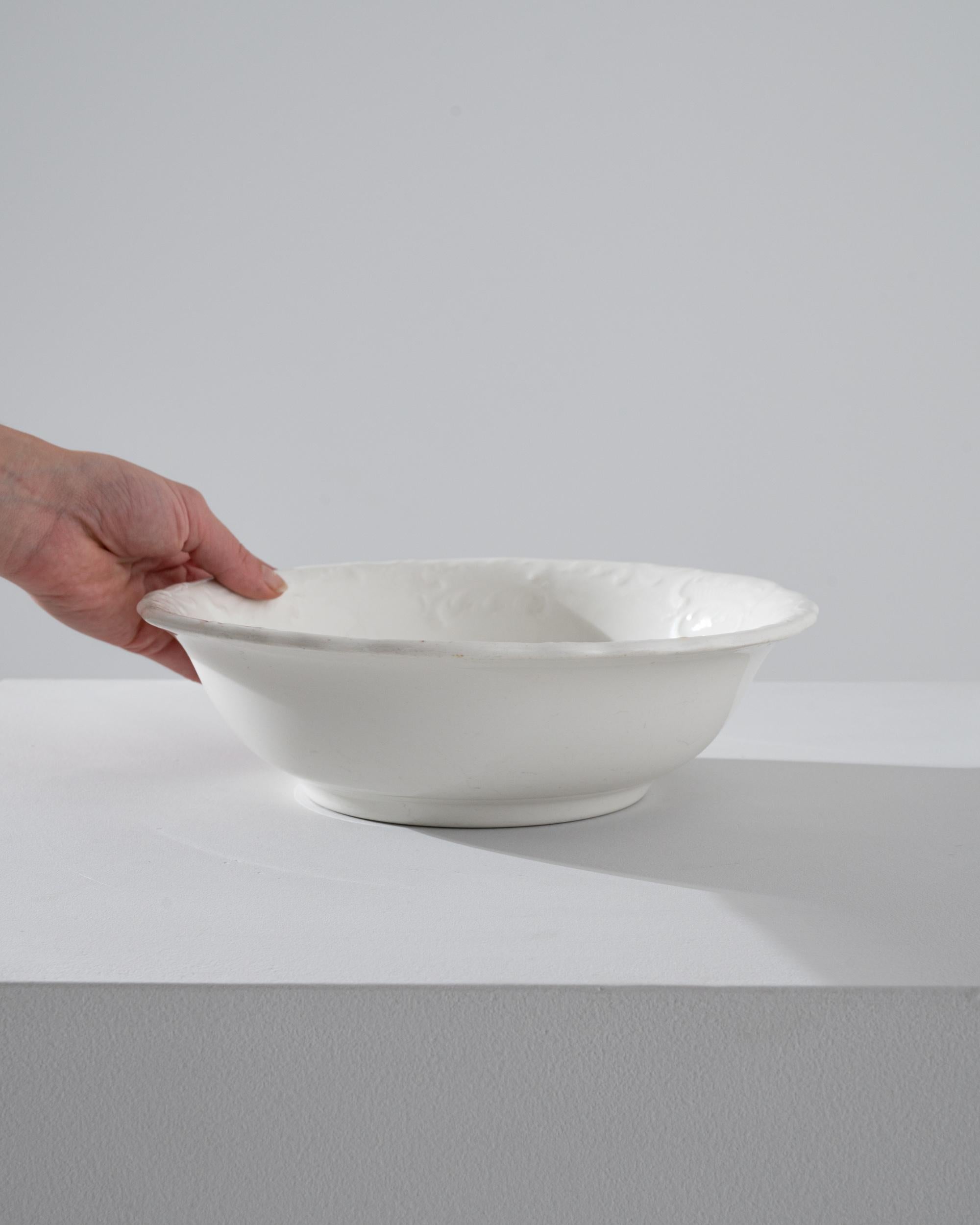 This 1900s Belgian porcelain bowl exudes a timeless charm with its understated elegance and classical simplicity. Its pristine white glaze and delicate scalloped edges speak of genteel afternoons and refined taste. The bowl's surface, adorned with