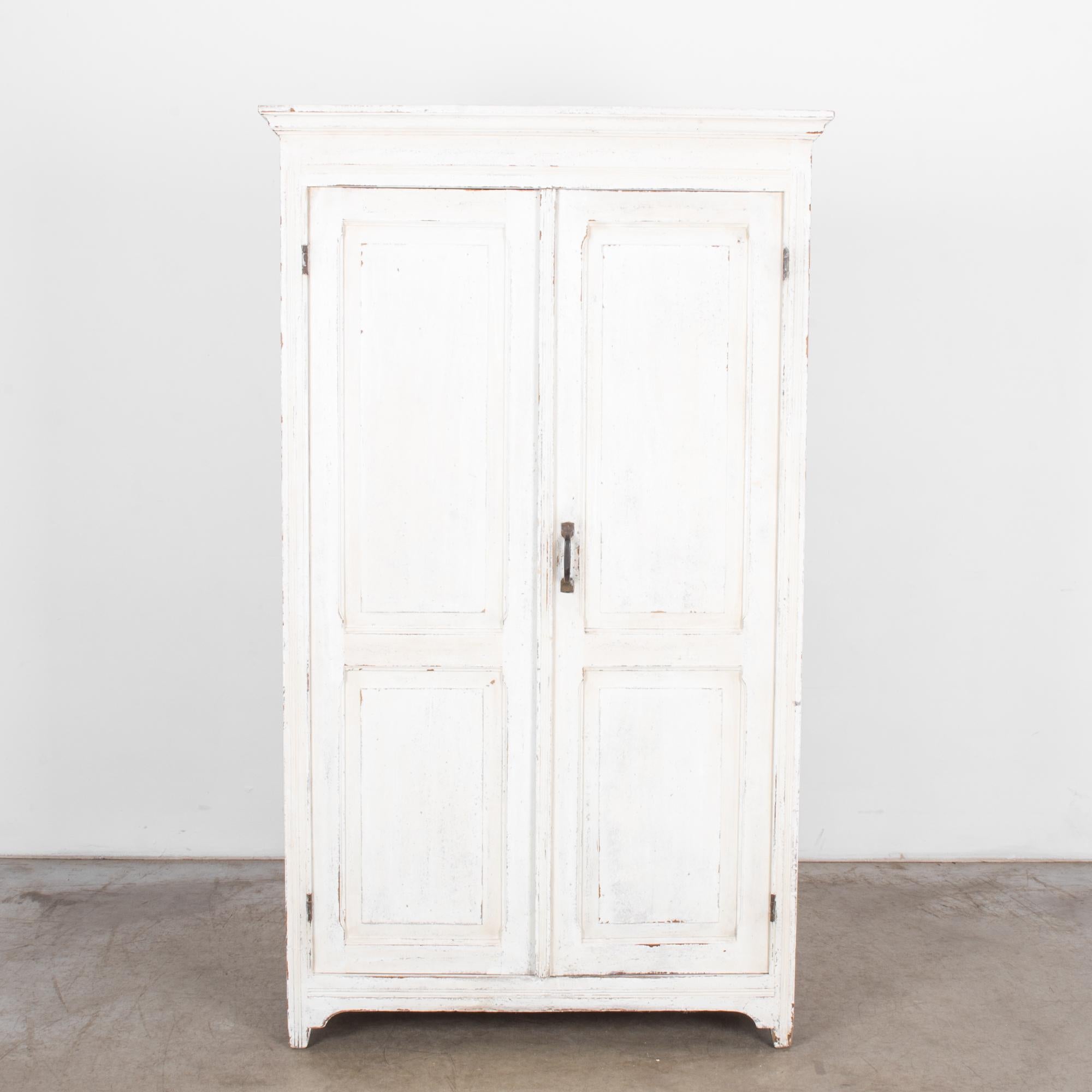An antique white wood patinated armoire from Belgium, circa 1900. Features wide double doors with recessed panels and curved brass handle, and a bifurcated interior with four shelves. White wash brings brightness and light to any affiliated interior.