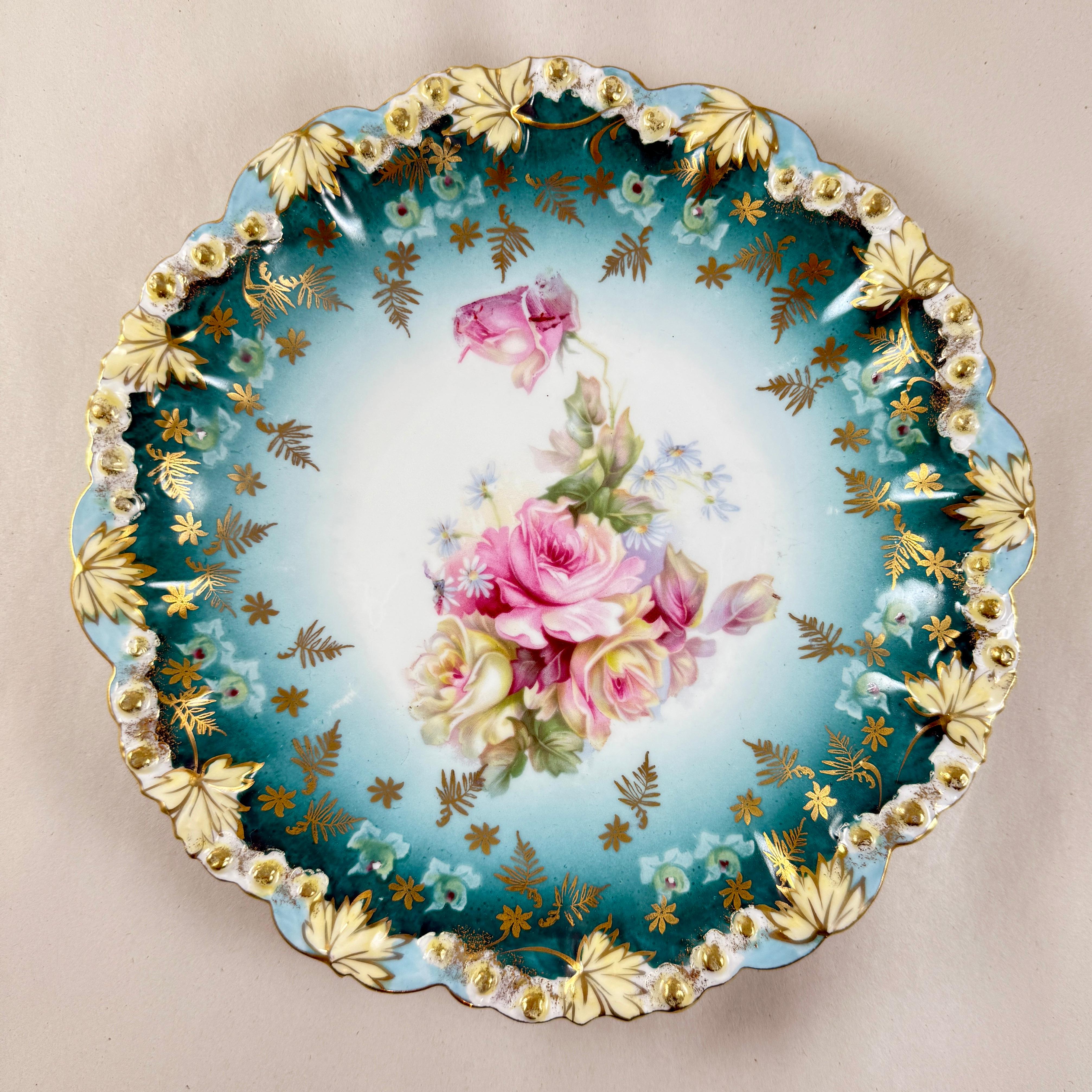 Ein tiefer Porzellanteller mit rosa Rosen und vergoldetem Blattrand, Bohemia, um 1900.

Ein satter tealblauer Grund, der zur Mitte hin verblasst und einen handgemalten Strauß aus rosa und gelben Rosen zeigt.

Eine komplexe innere Bordüre mit einer