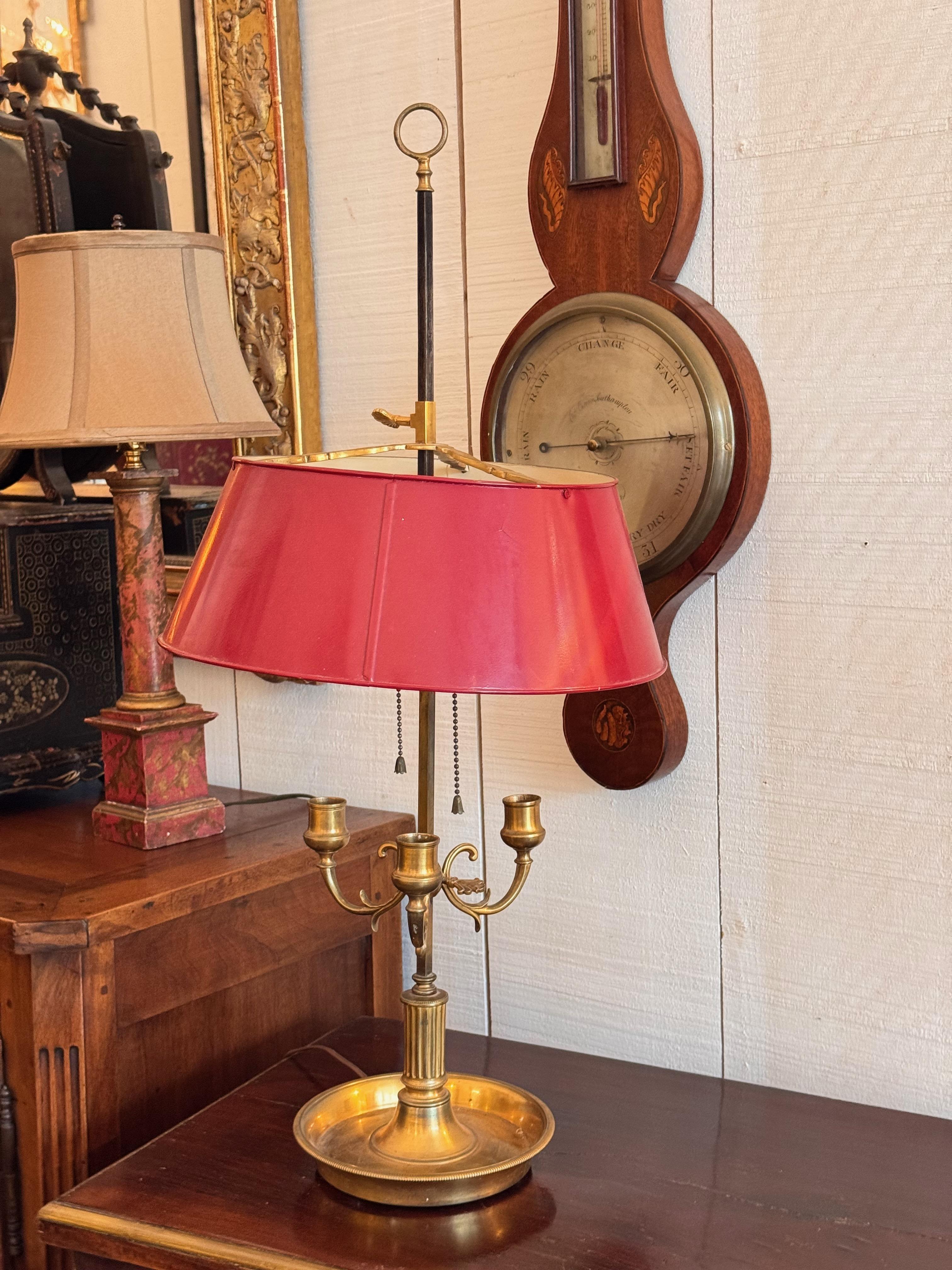 Lampada bouillotte classica in ottone con tre bracci curvi, una colonna centrale scanalata e una base rotonda a goccia. Il paralume in tole rosse è regolabile in altezza lungo l'asta centrale, un design originariamente pensato per schermare la luce