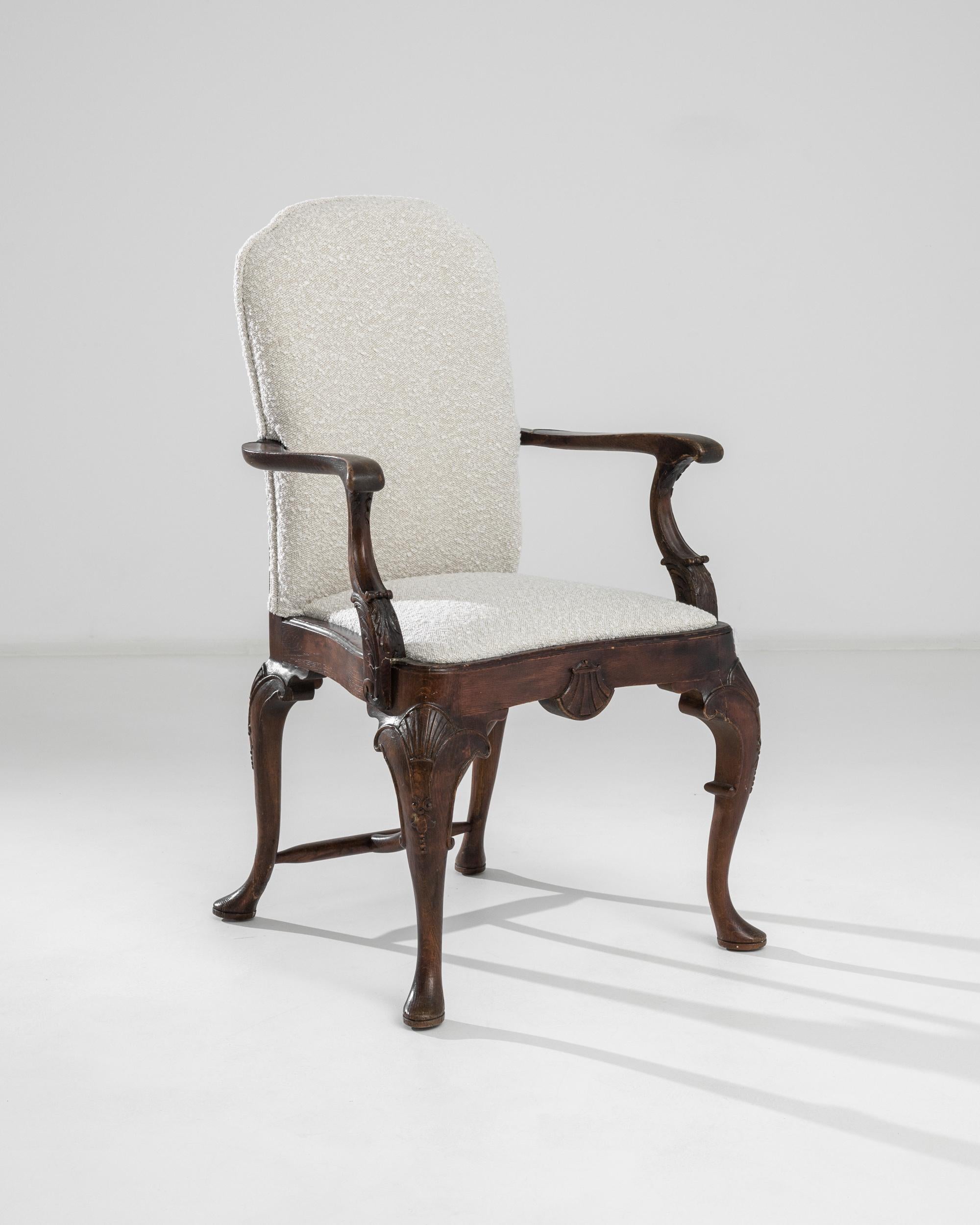 This upholstered wooden armchair from turn of the century Britain offers a distinguished yet inviting seat. An upright seat atop a graceful cabriole base creates a throne-like silhouette. The burnished brown patina of the oak accentuates the rococo