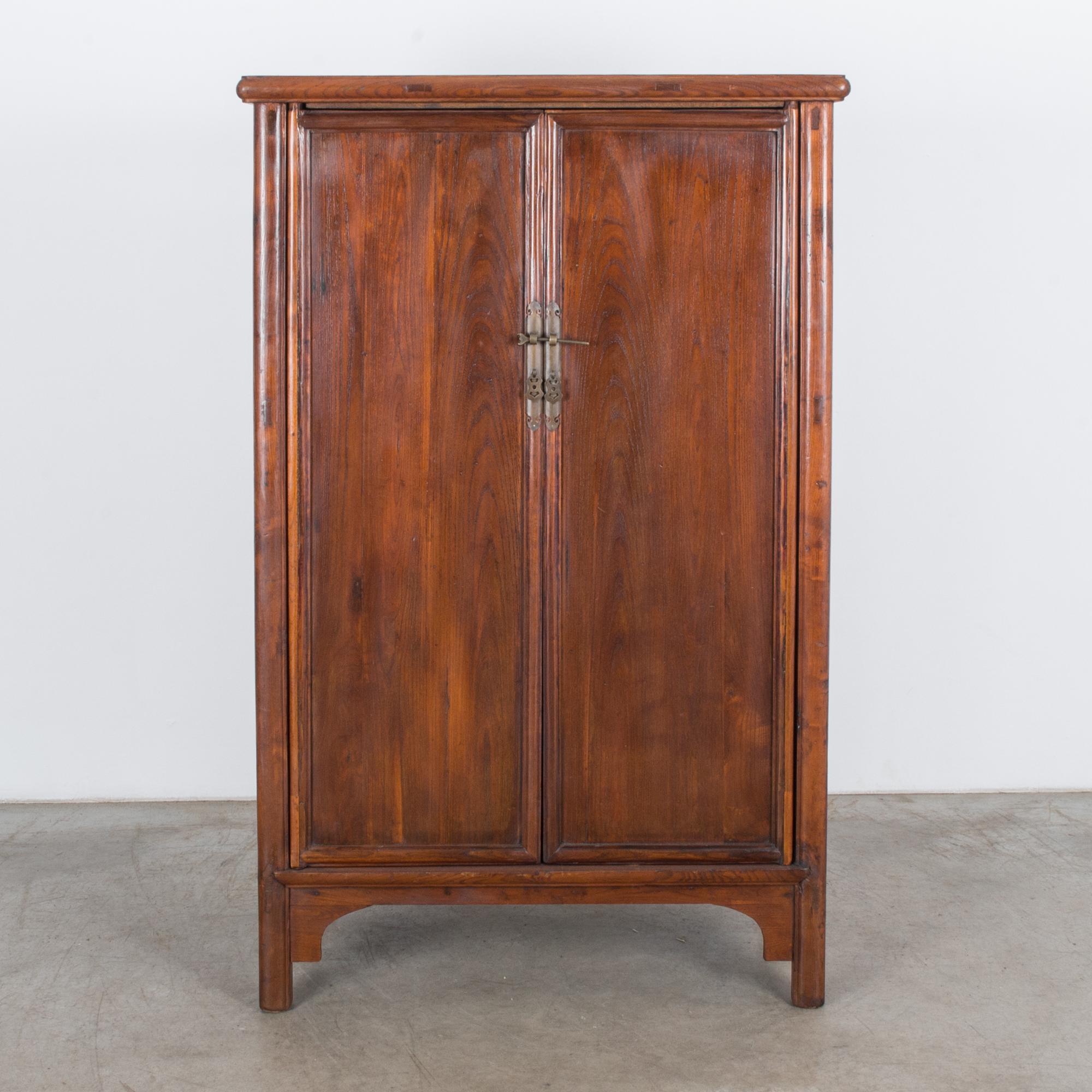 This wooden armoire was made in China, circa 1900. The double doors open to reveal a spacious interior with a shelf and two drawers. A decorative locking pin secures the doors, and it is slightly raised on four legs. With its lovely patina and rich