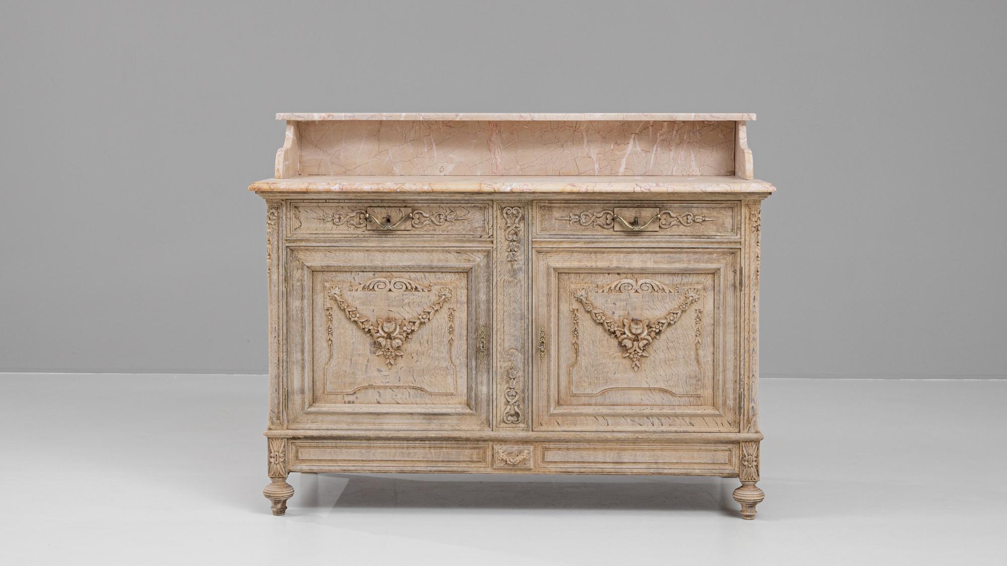 This stunning French buffet from the early 1900s exemplifies timeless elegance with its bleached oak finish and exquisite marble top. The piece features intricate carvings, including floral and garland motifs on its doors, which bring a touch of