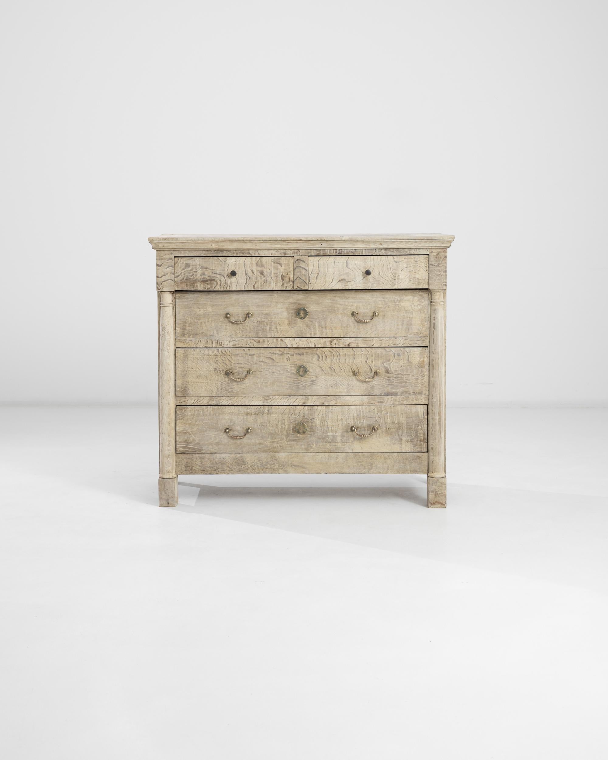 Built in France circa 1900, this vintage oak chest of drawers presents a simple yet refined constitution: two shallow drawers rest atop three ample compartments adorned by gilded bail pulls and decorative locks. Neoclassical elements such as a