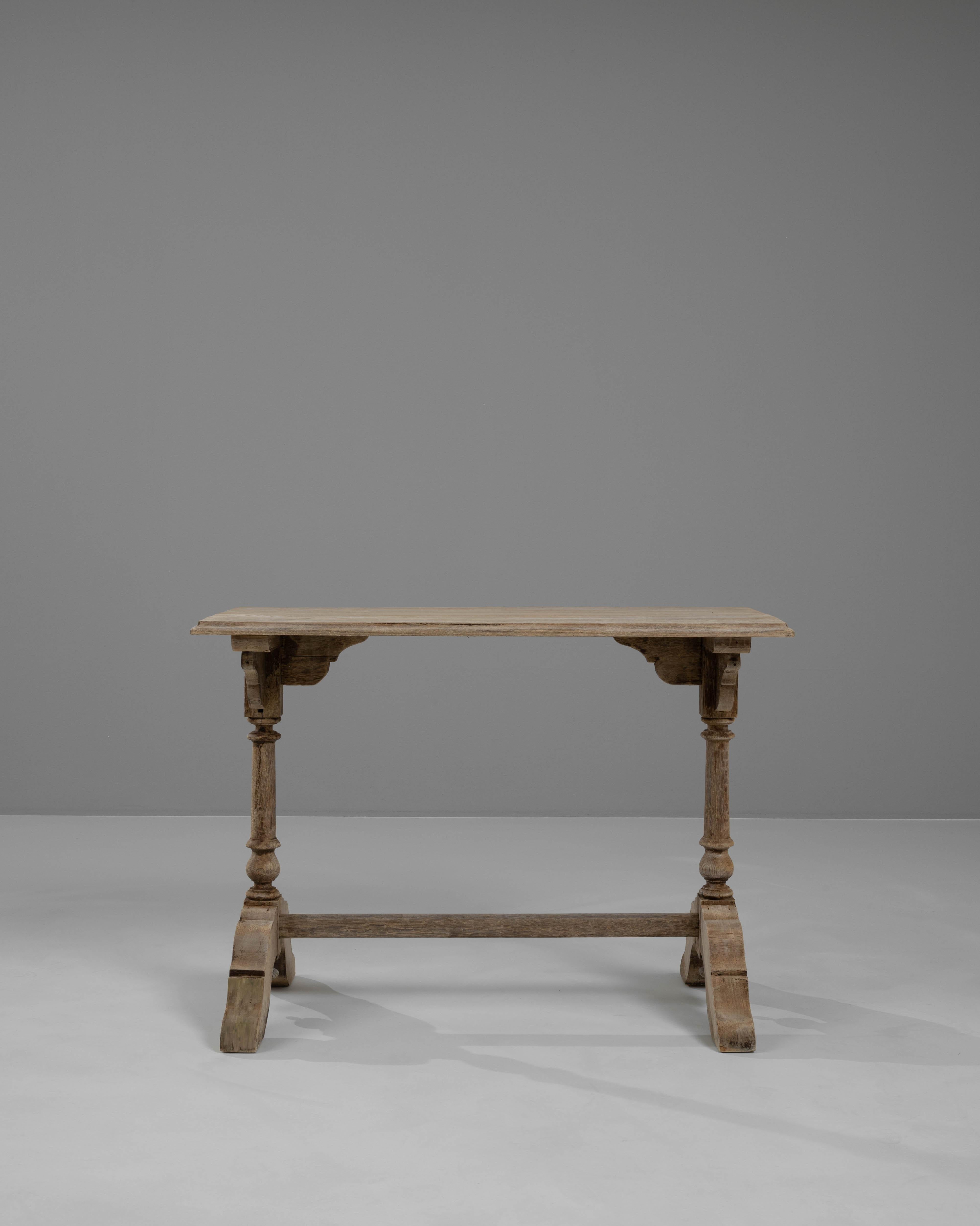 This 1900s French bleached oak side table is a masterpiece of antique design, exuding old-world charm and sophisticated elegance. The table features a unique dual pedestal base, each ending in ornately carved feet with original casters for easy