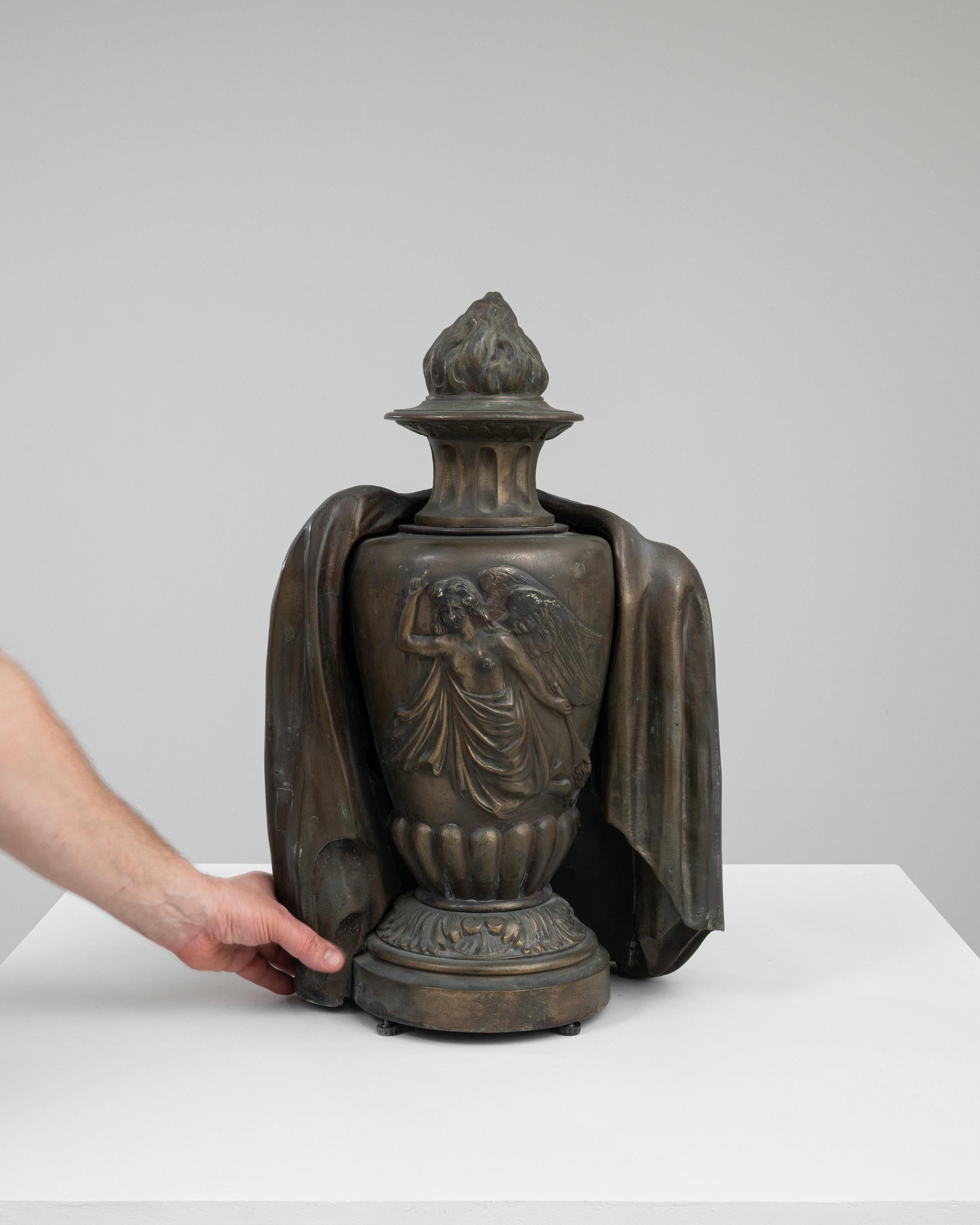 Cette captivante urne en bronze française des années 1900 est une relique exquise qui incarne l'art et la mystique de son époque. Drapée des plis gracieux d'une robe classique, l'urne est ornée de la figure sereine et sculptée d'un ange, apportant